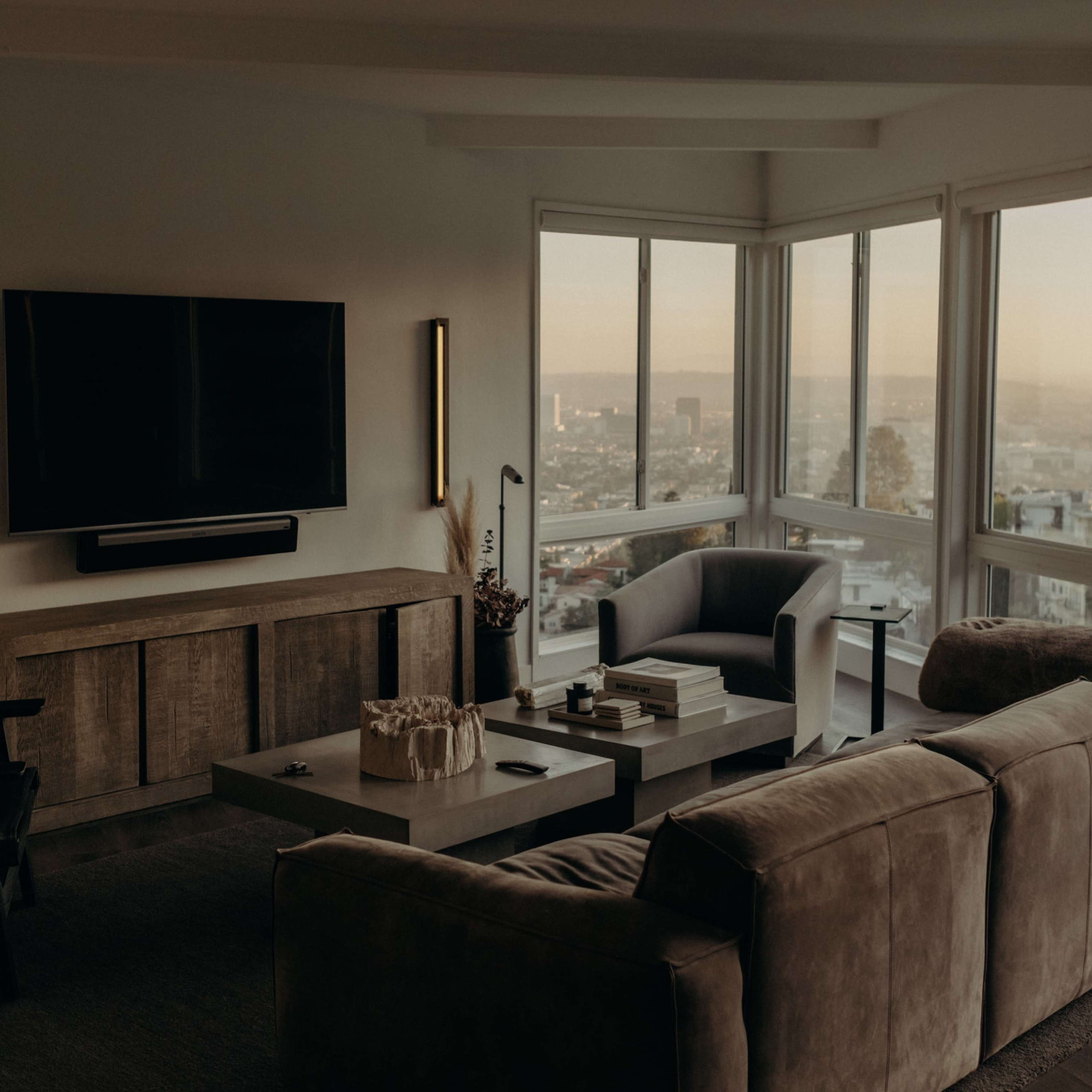 A modern living room features a large window with a view of the city, a sectional sofa, and a mounted TV on a sleek cabinet.