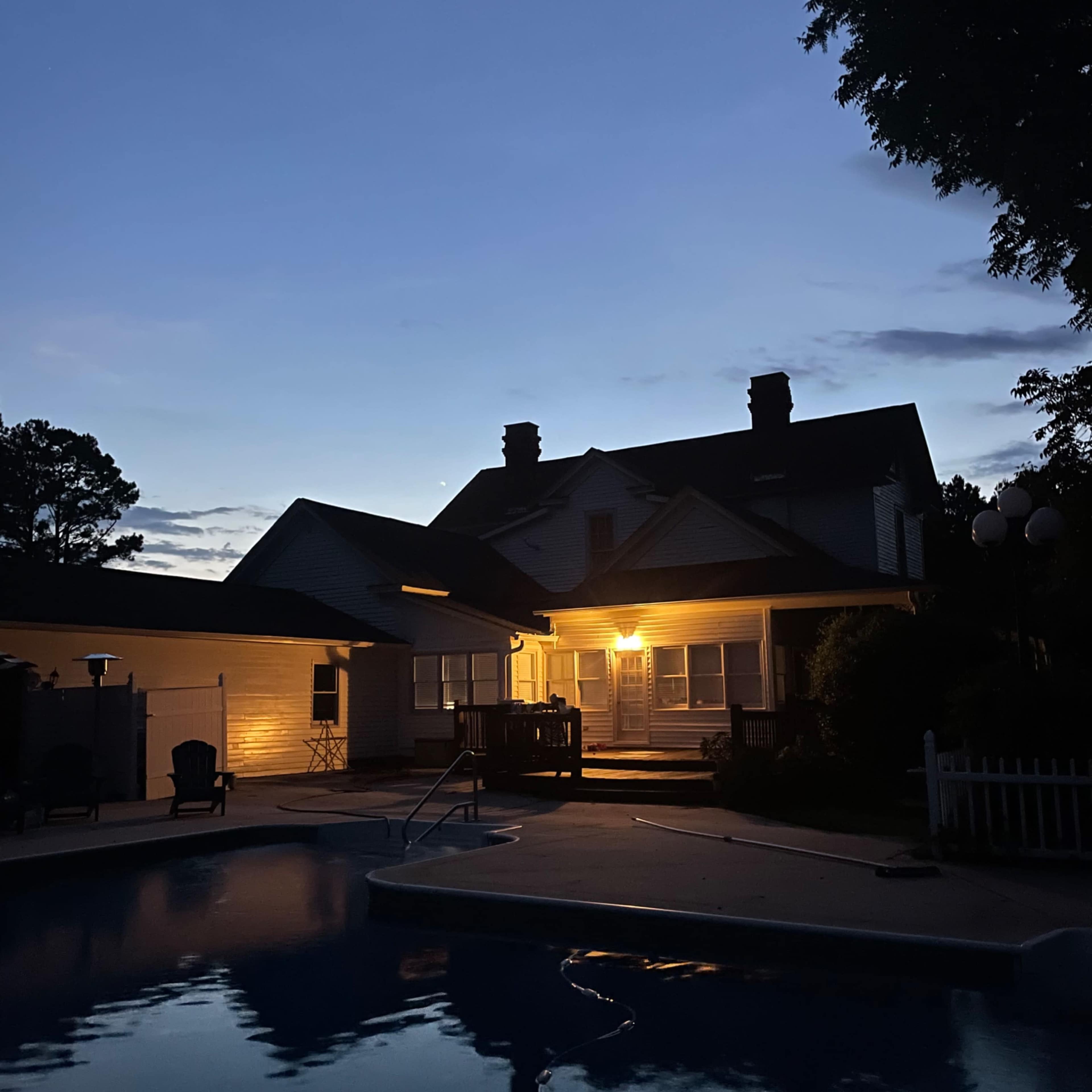 A two-story house with a lit porch stands beside a swimming pool at dusk.