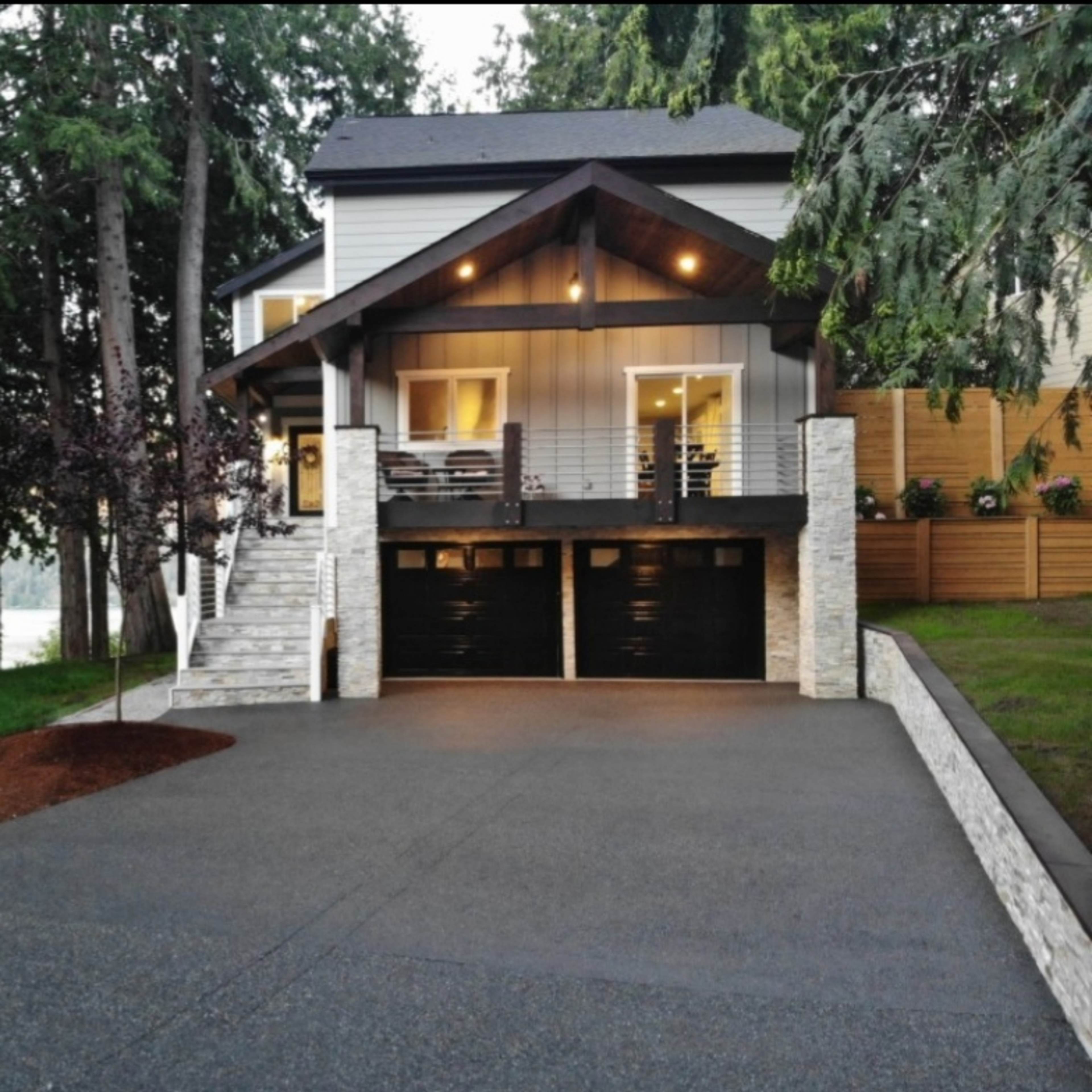 A modern house with a large front porch, two black garage doors, and a landscaped driveway is surrounded by trees.