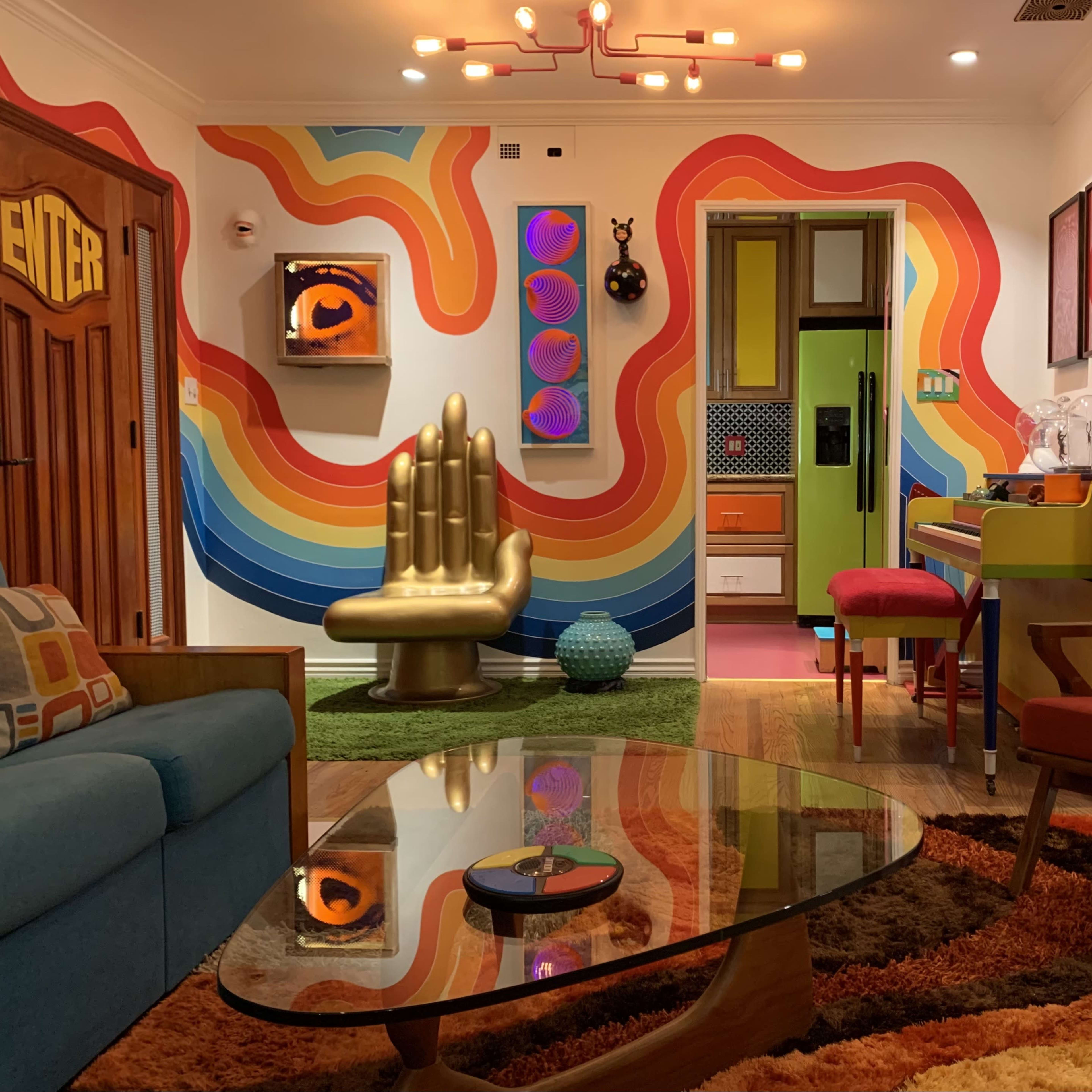 The room features a colorful retro design with a rainbow mural on the wall, a gold hand sculpture, and modern furniture including a glass coffee table and a bright red armchair.