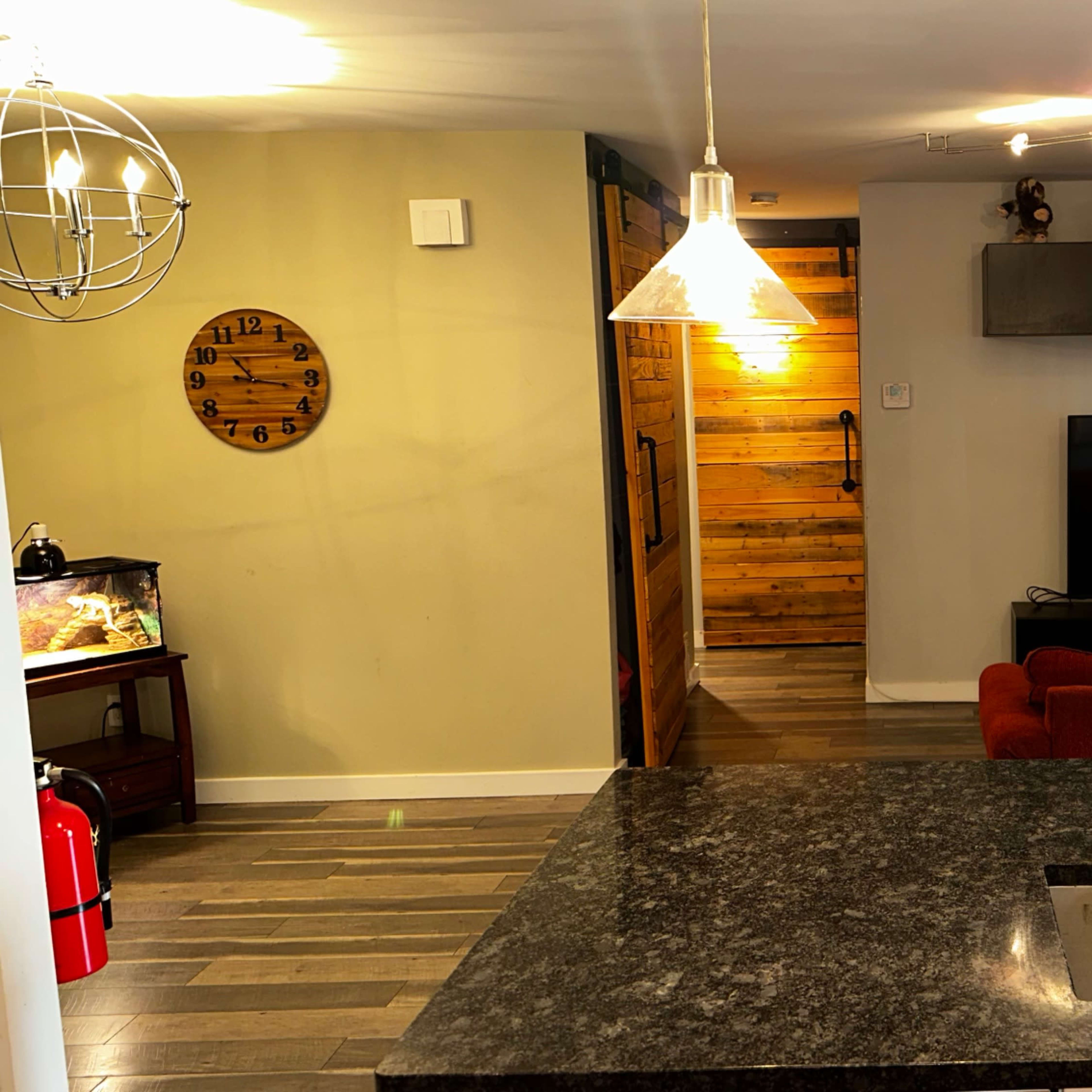 A modern kitchen with a dark granite countertop, a dining area in the foreground, and a living room featuring a television and a guitar hanging on the wall.