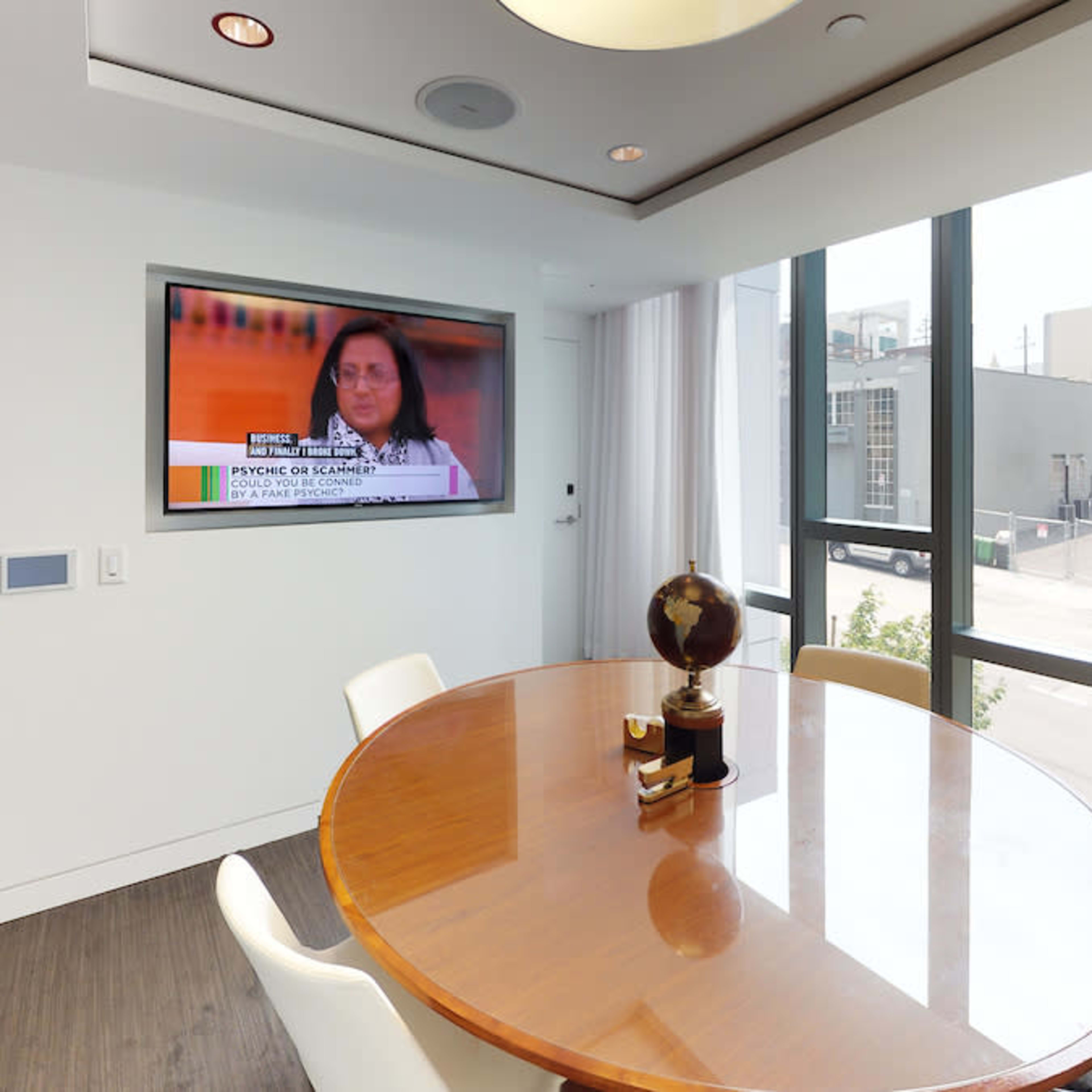 A round wooden table with chairs is seen in a well-lit room featuring a large television displaying a news segment.