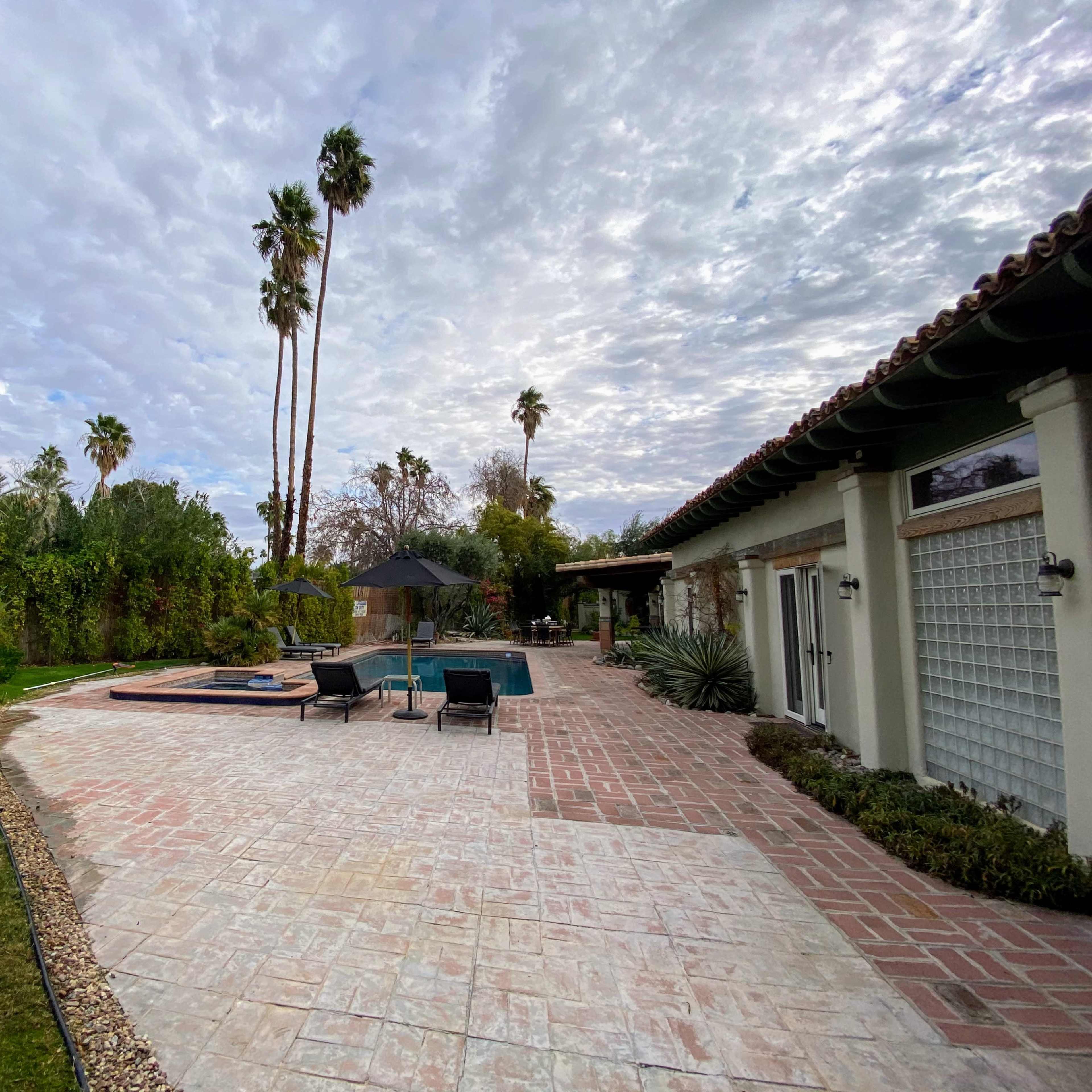 A backyard area featuring a swimming pool, seating by the pool, and palm trees under a cloudy sky.