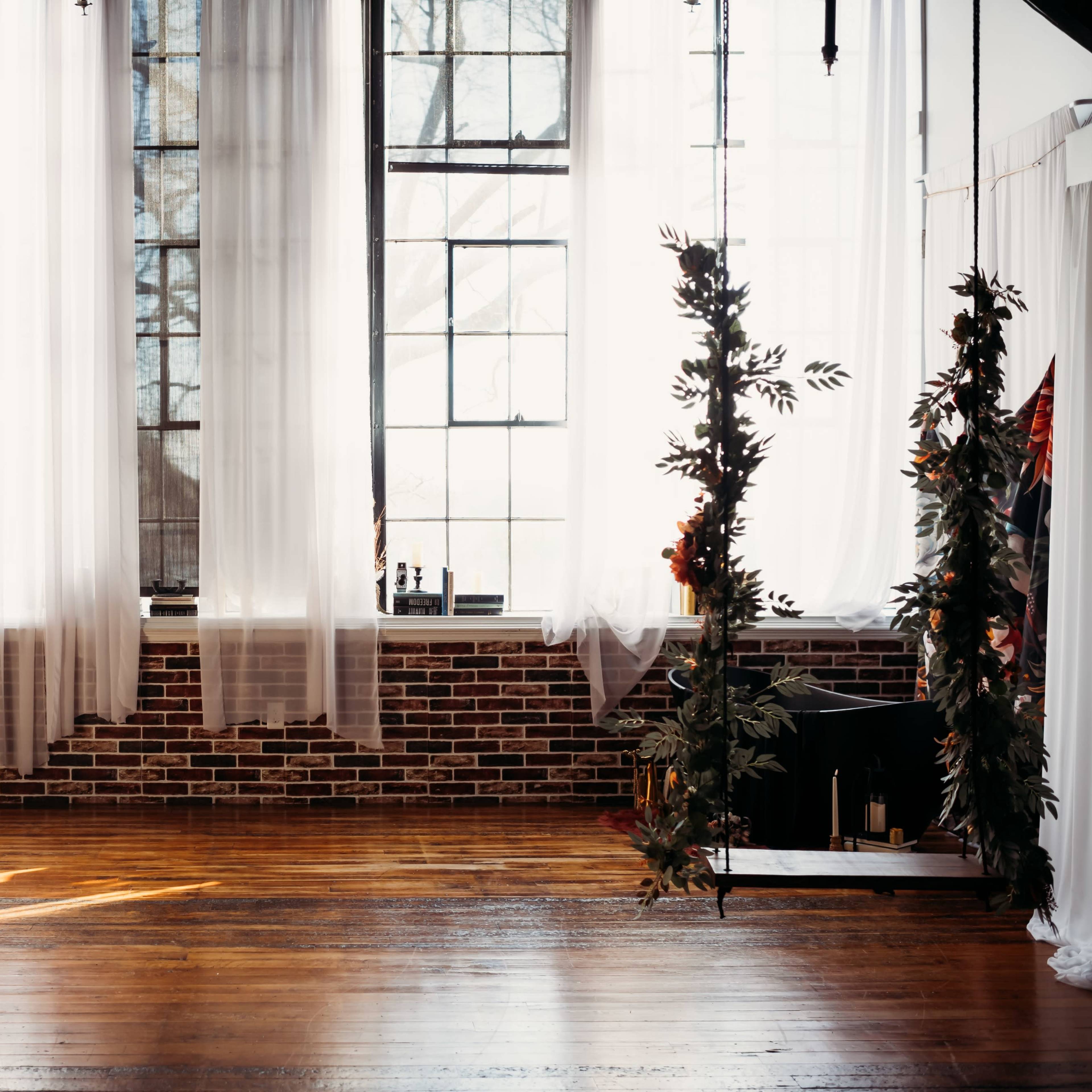 A spacious interior featuring large windows with sheer curtains, exposed brick walls, and wooden floors, with a floral backdrop and a hanging plant display.