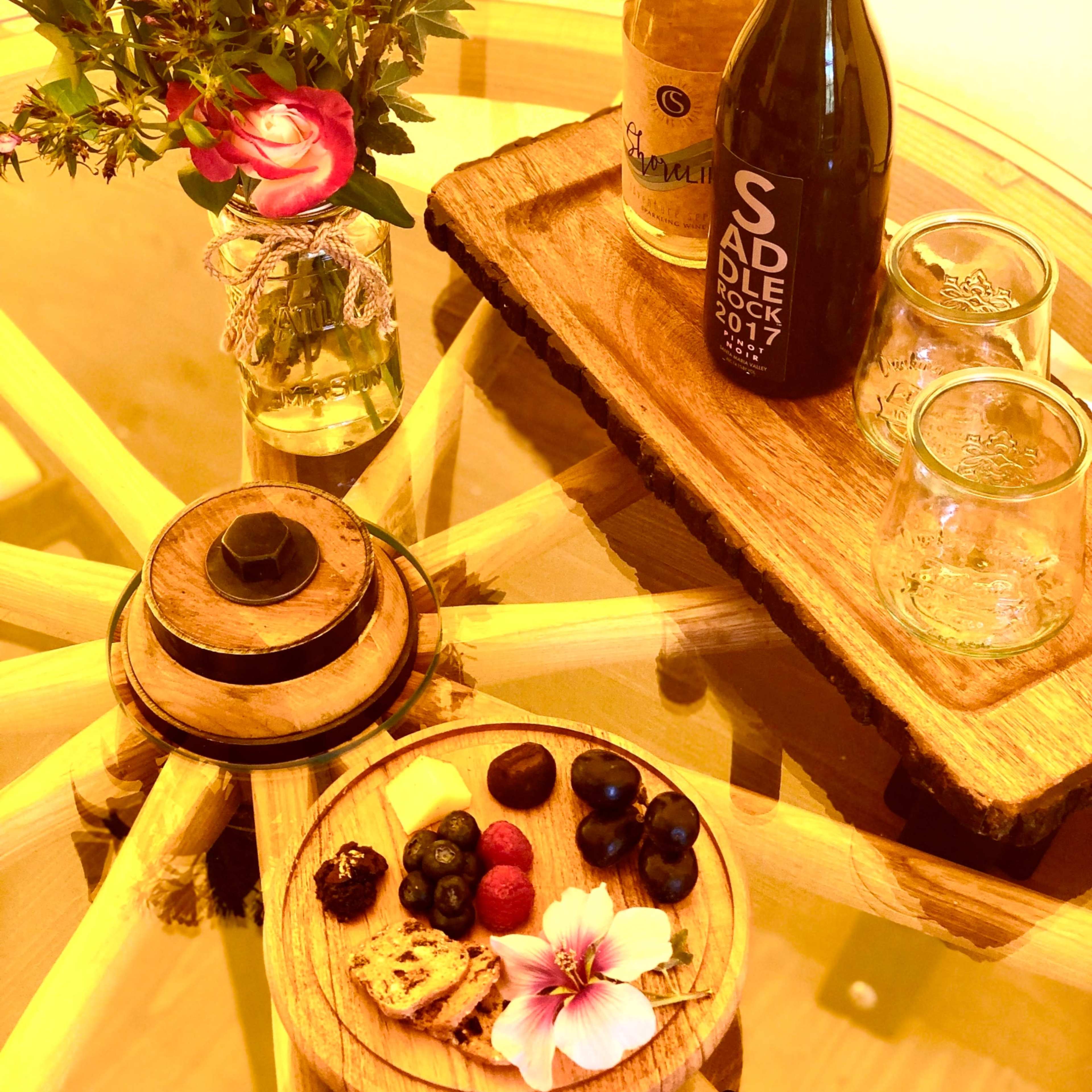 A wooden table displays a bottle of white wine, a bottle of red wine, two glasses, a plate of assorted fruits and cookies, and a vase of flowers.