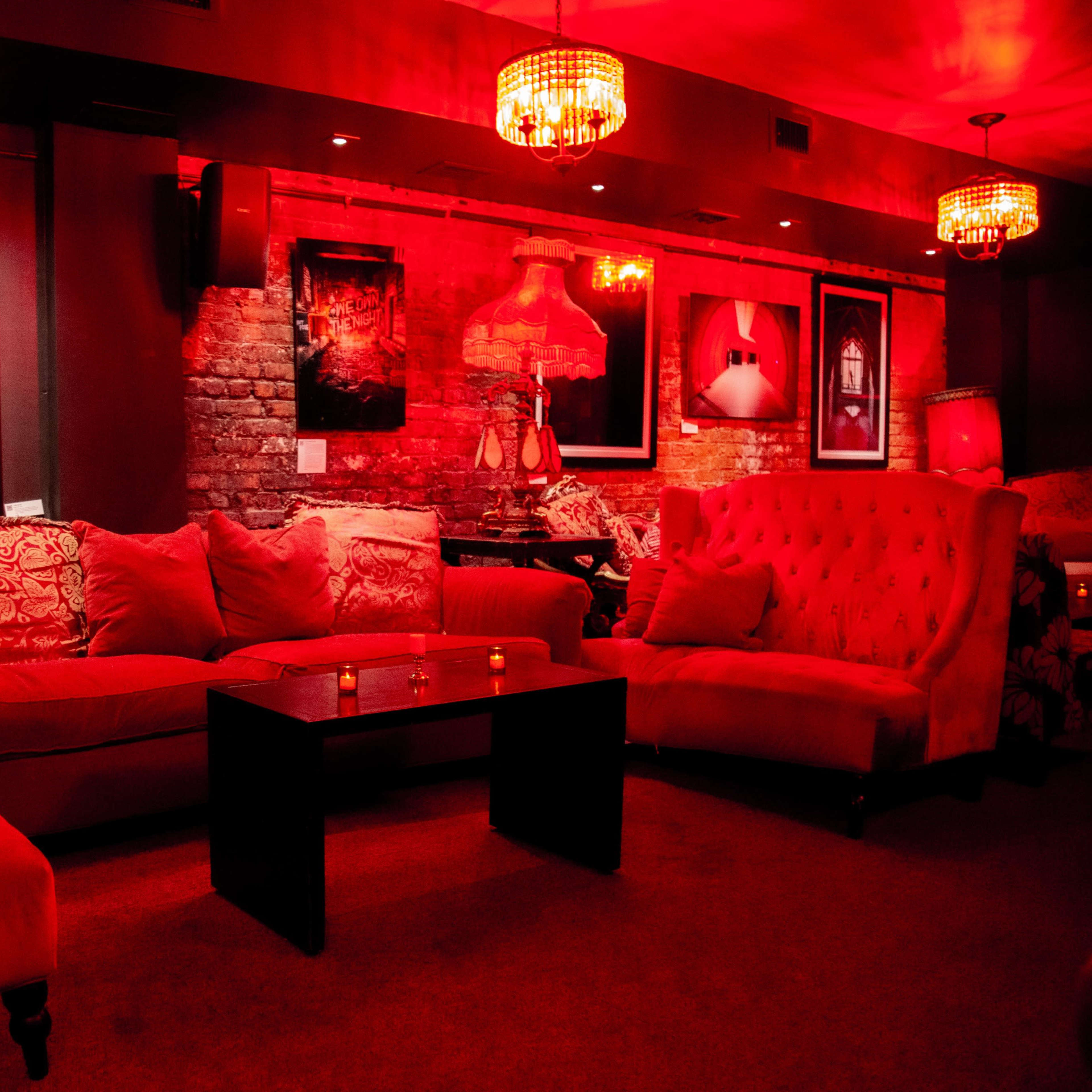 A dimly lit lounge area with red lighting, featuring plush red sofas, a dark coffee table, and framed artwork on the walls.