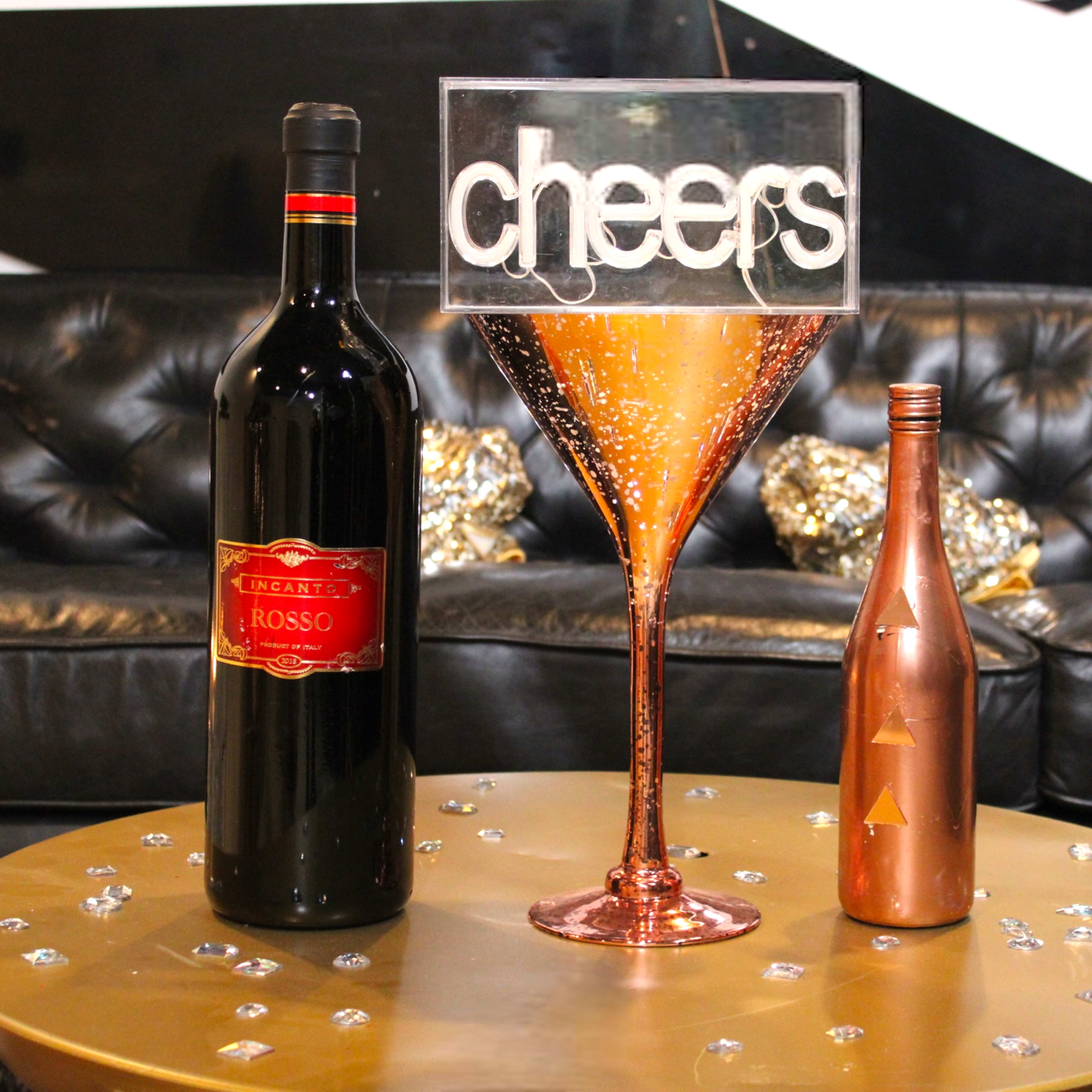 A large decorative martini glass filled with glimmering liquid stands on a gold table, surrounded by a bottle of red wine and a smaller bottle, against a backdrop of a black leather couch and metallic decor.