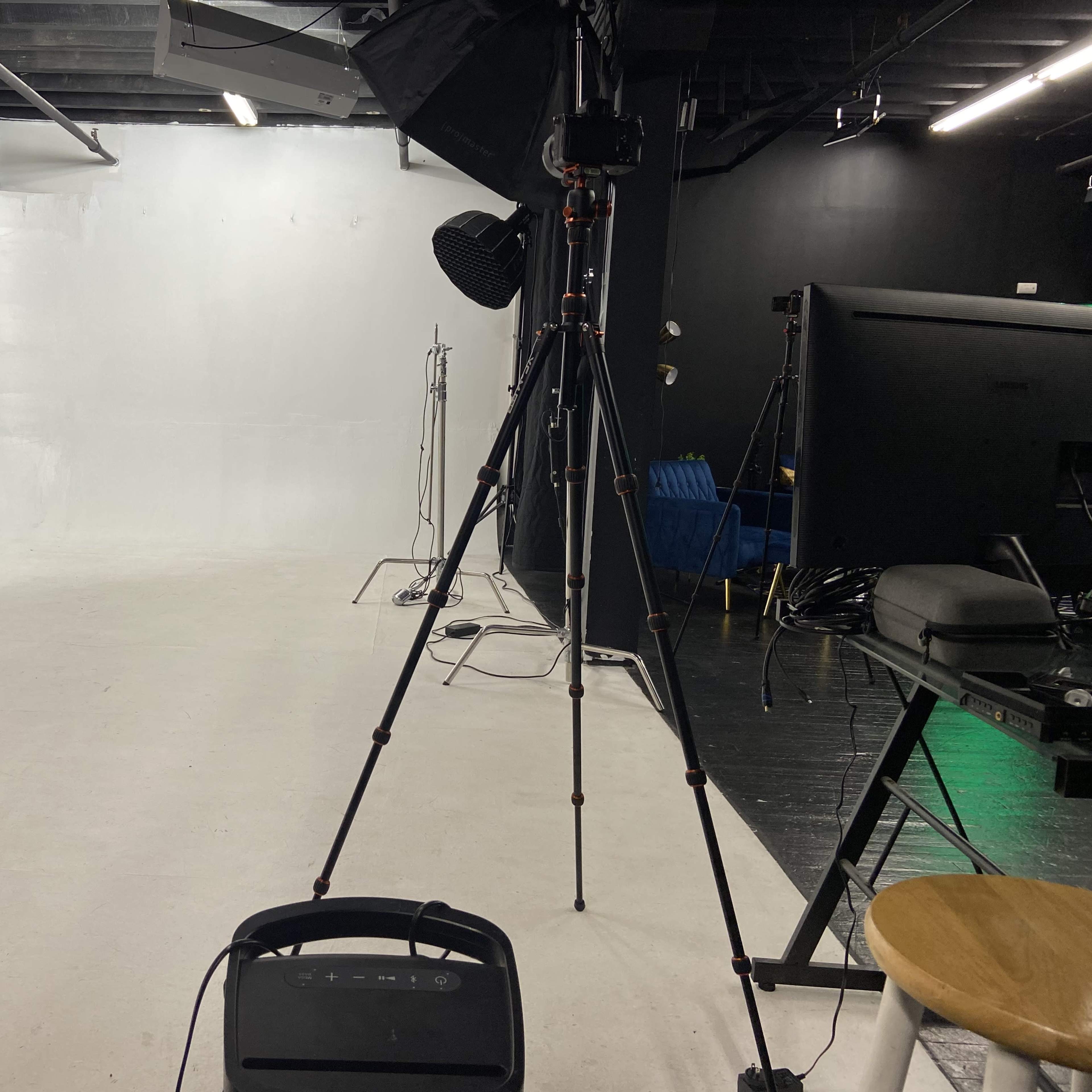 A photography studio with a white backdrop, equipment including a tripod, lighting setup, and a monitor.