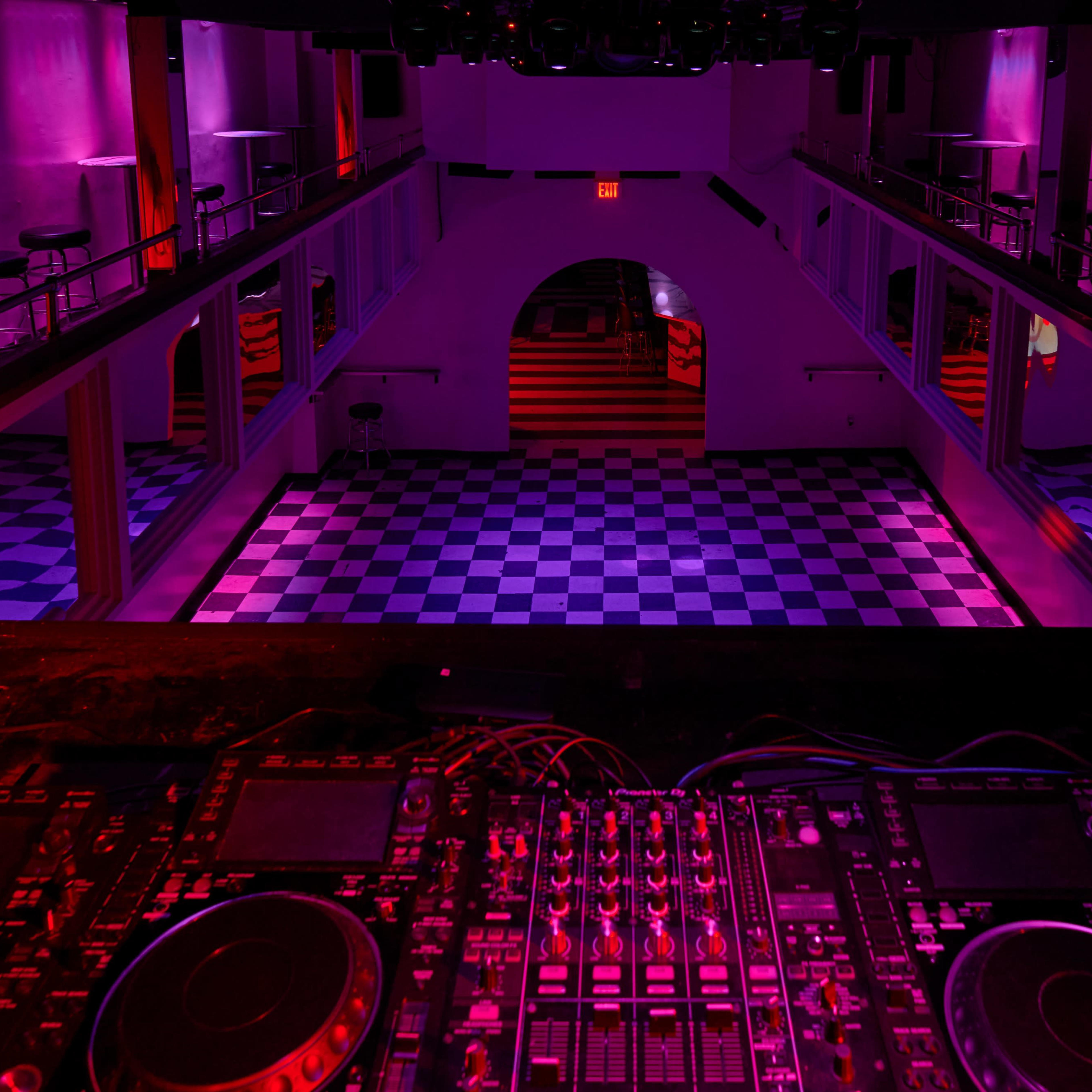 An empty nightclub from a high vantage point, featuring a checkered dance floor and illuminated walls in shades of pink and purple.
