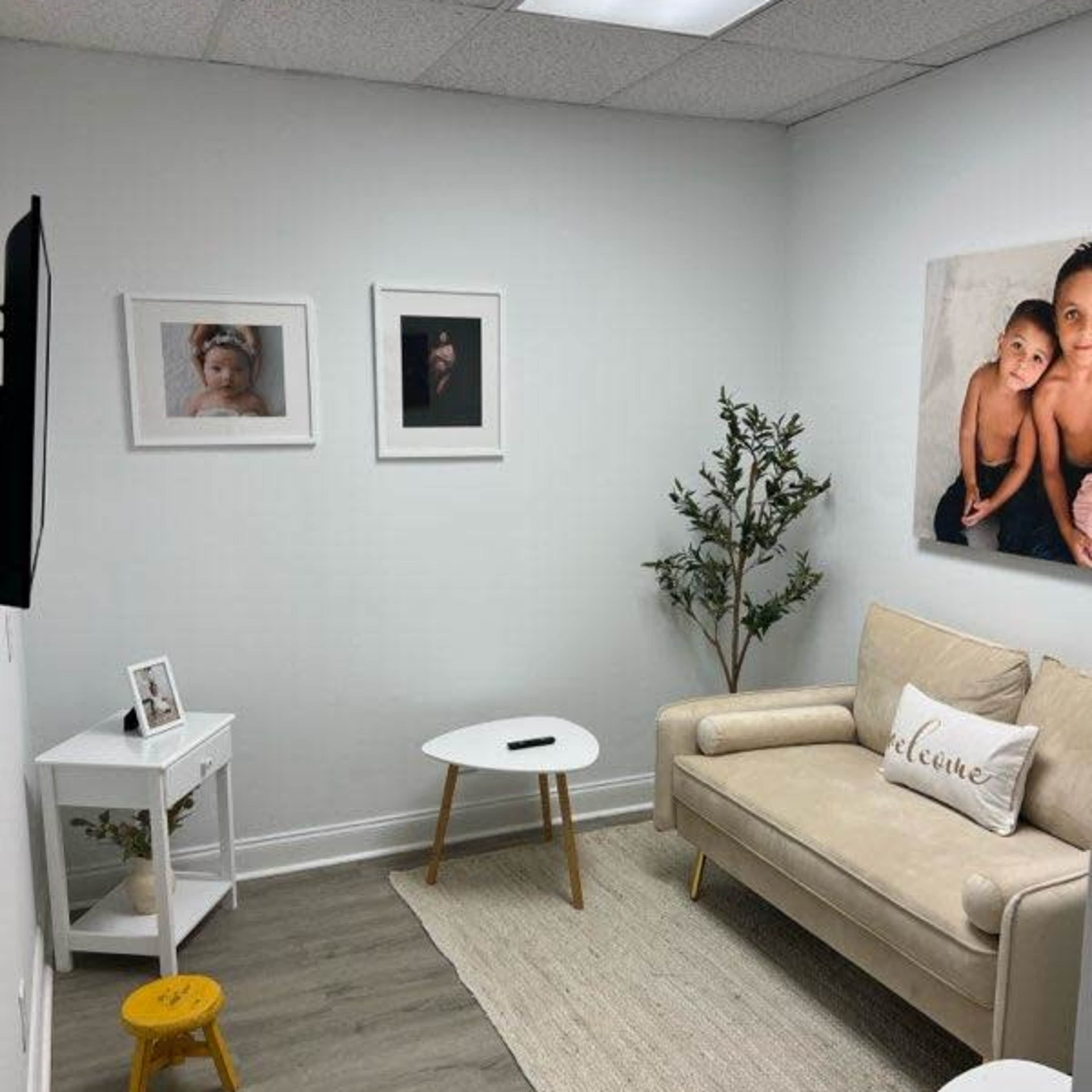 A small, neatly arranged waiting room featuring a beige couch, decorative photos on the walls, a small table with a plant, and a light-colored rug.