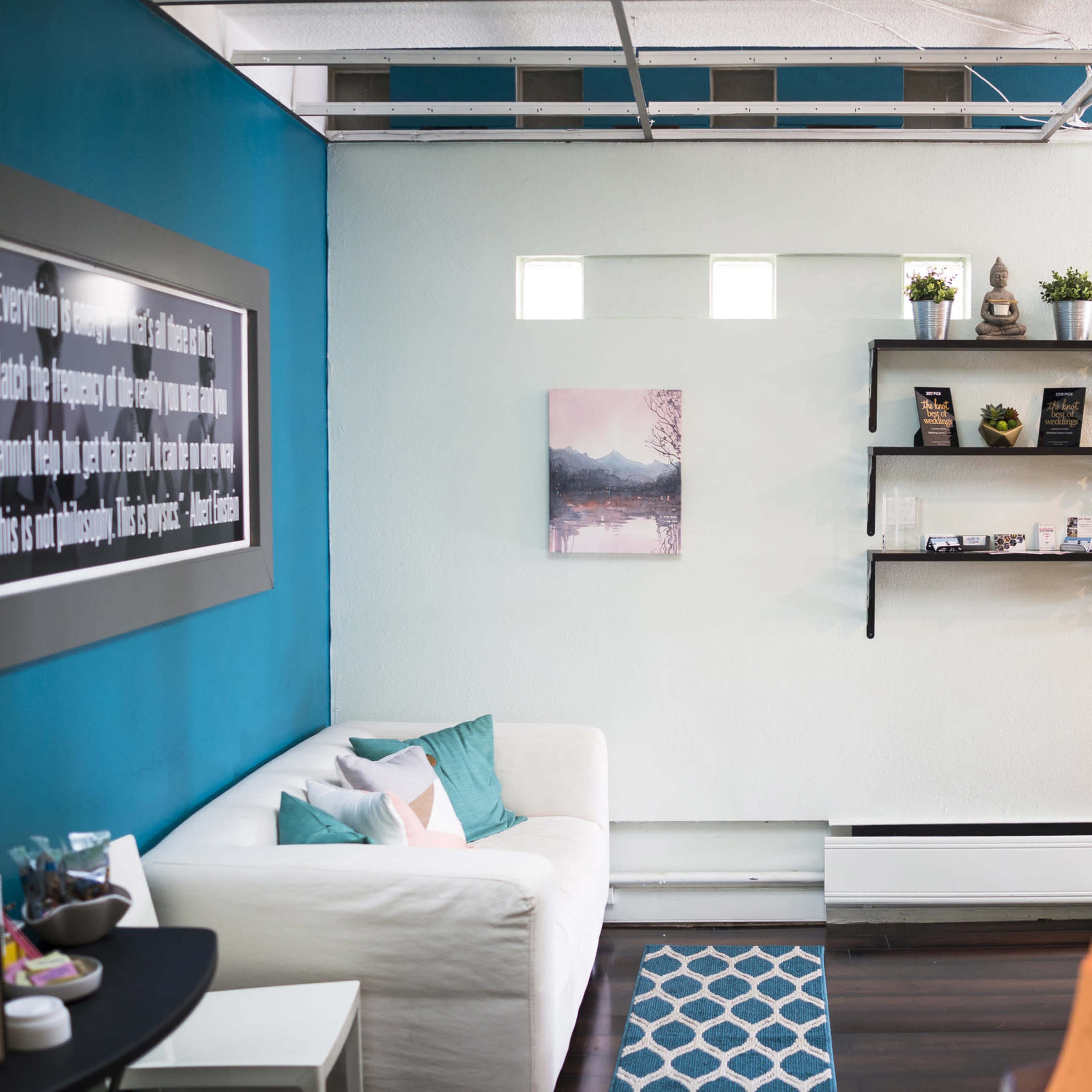 A modern, minimalist interior with a white sofa, a teal accent wall, and decorative shelves displaying plants and items.