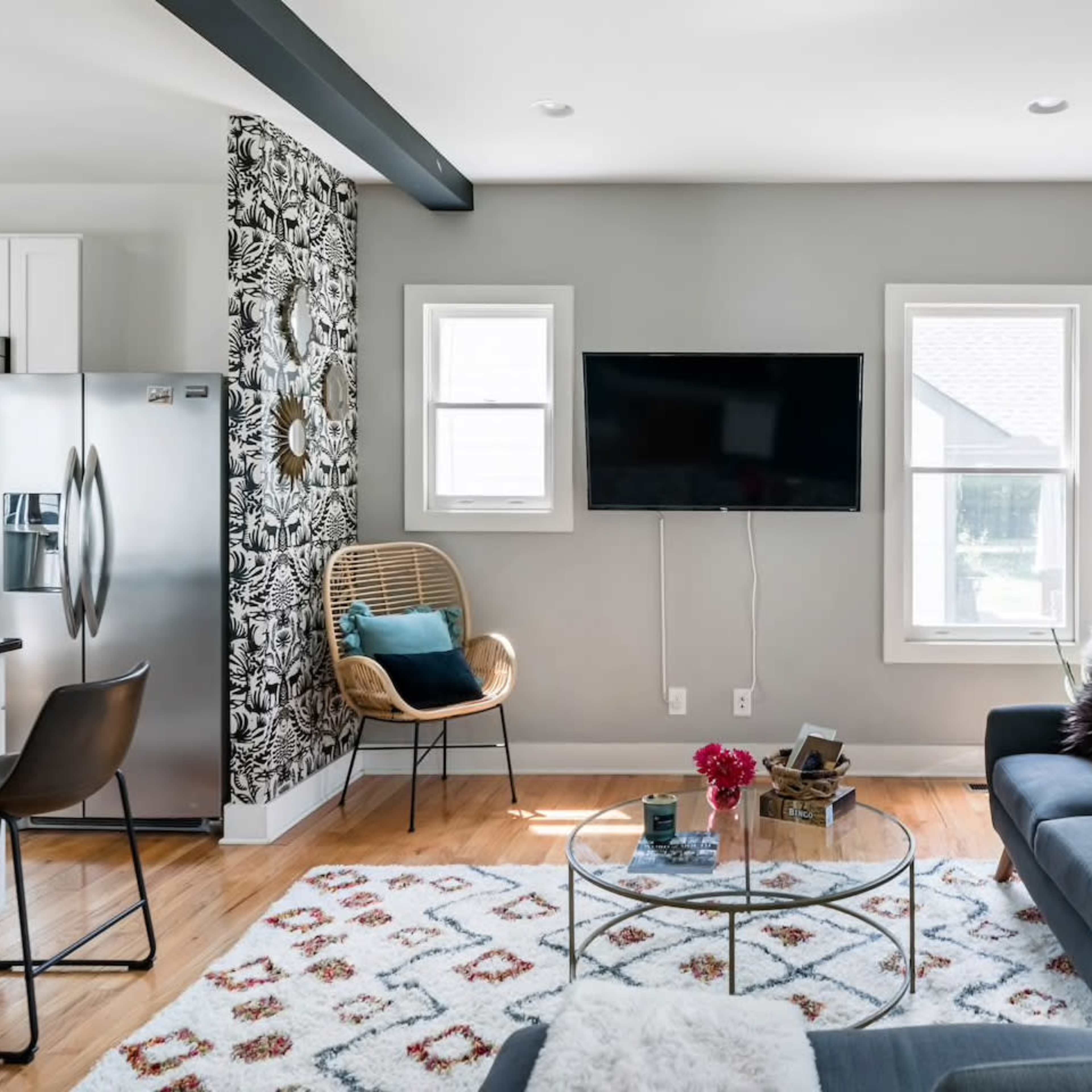 A modern living room features a gray sofa, a glass coffee table, a television mounted on the wall, and a patterned area rug.