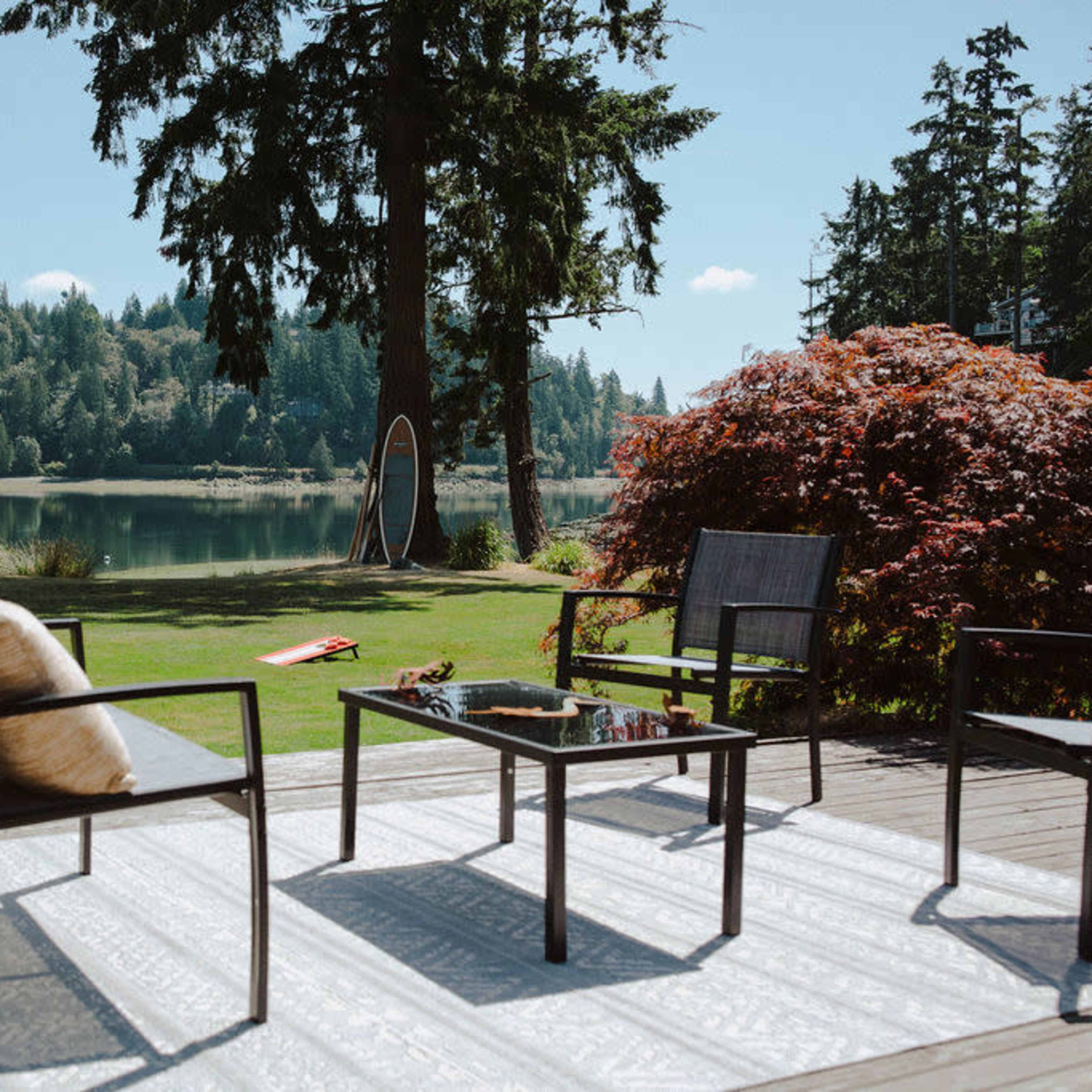 A wood deck features a set of black chairs and a table, overlooking a calm lake surrounded by trees and a colorful shrub.