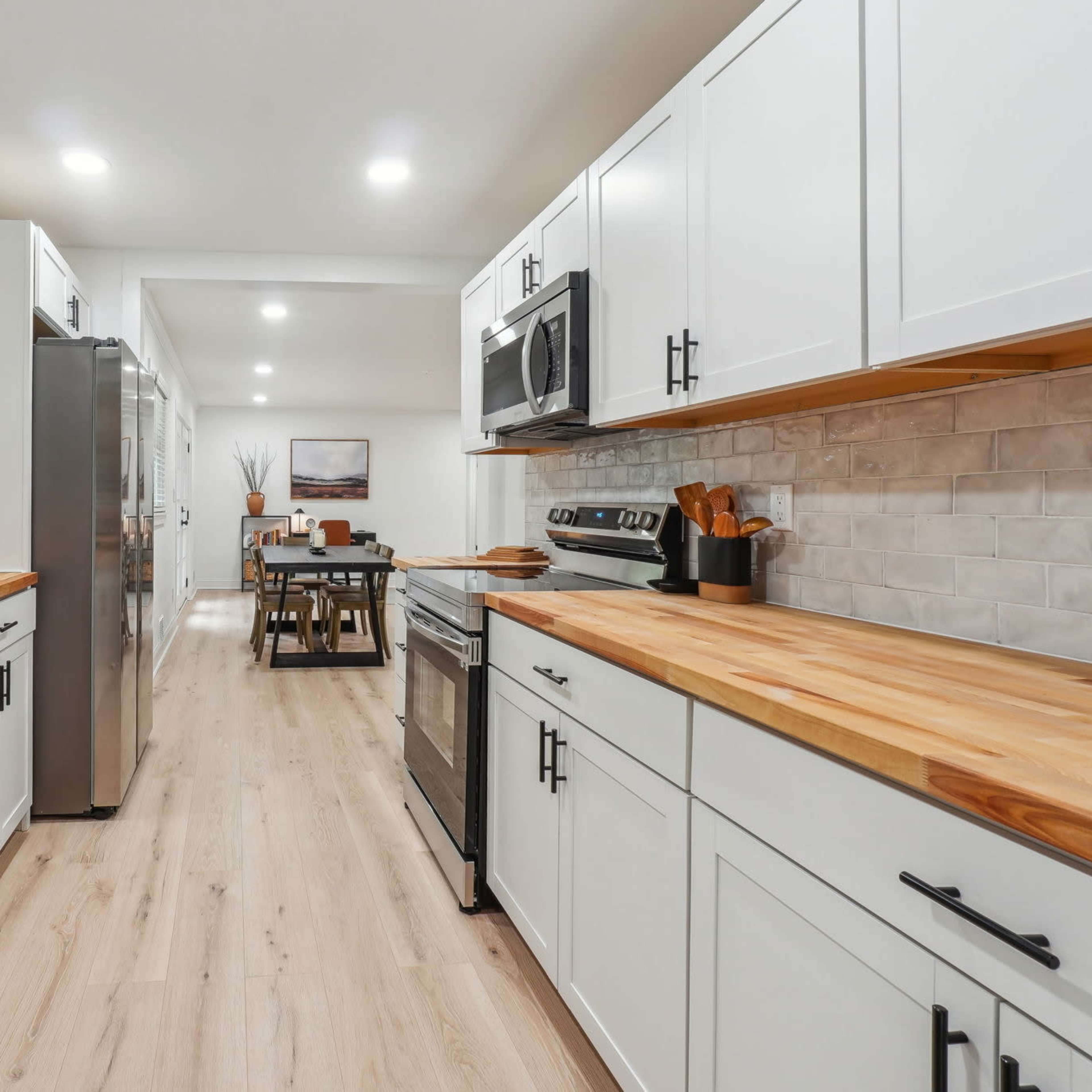 A modern kitchen with white cabinets, stainless steel appliances, and wooden countertops, leading into a dining area in the background.