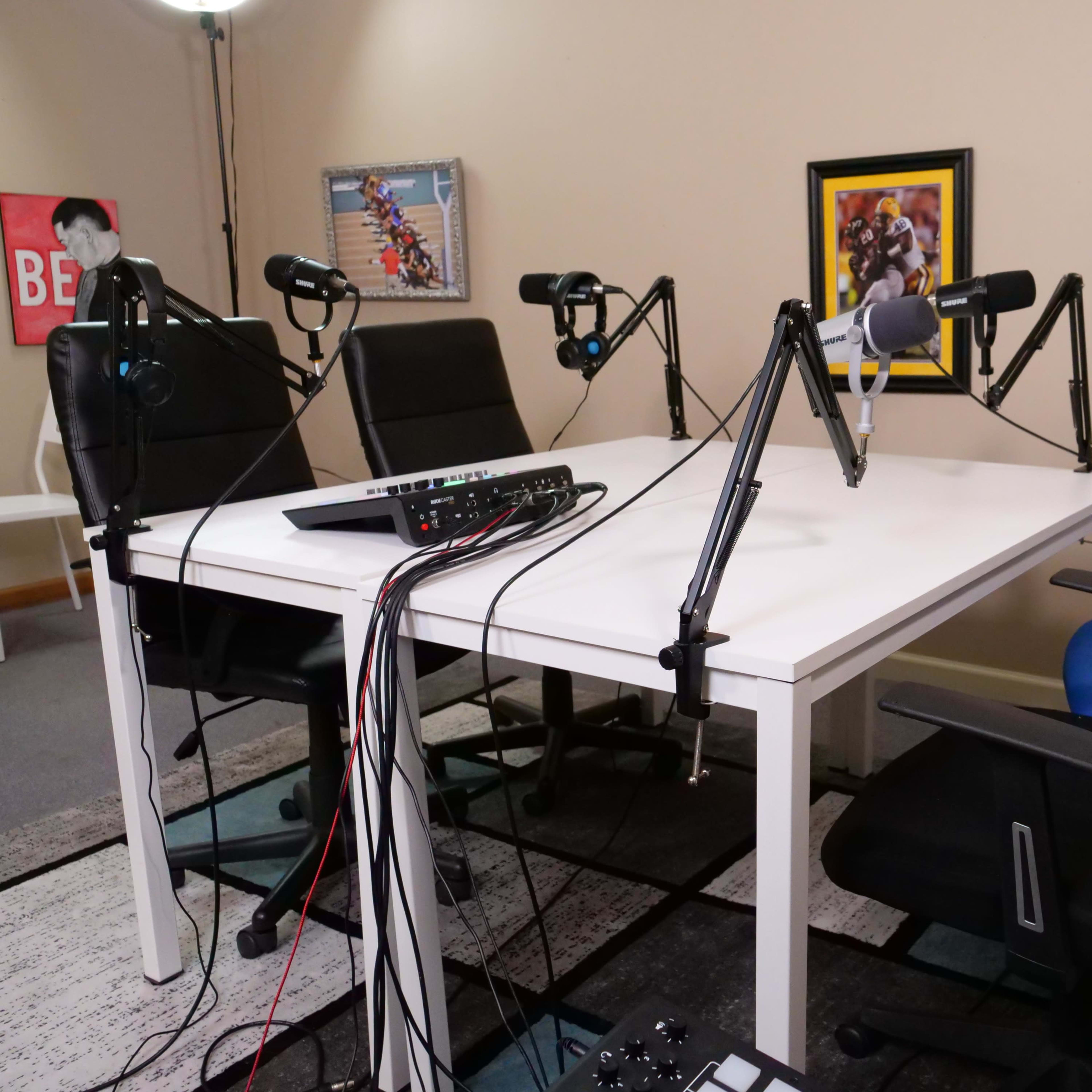 A podcast recording setup with a white table, multiple microphones on stands, and four chairs of varying colors arranged around the table.