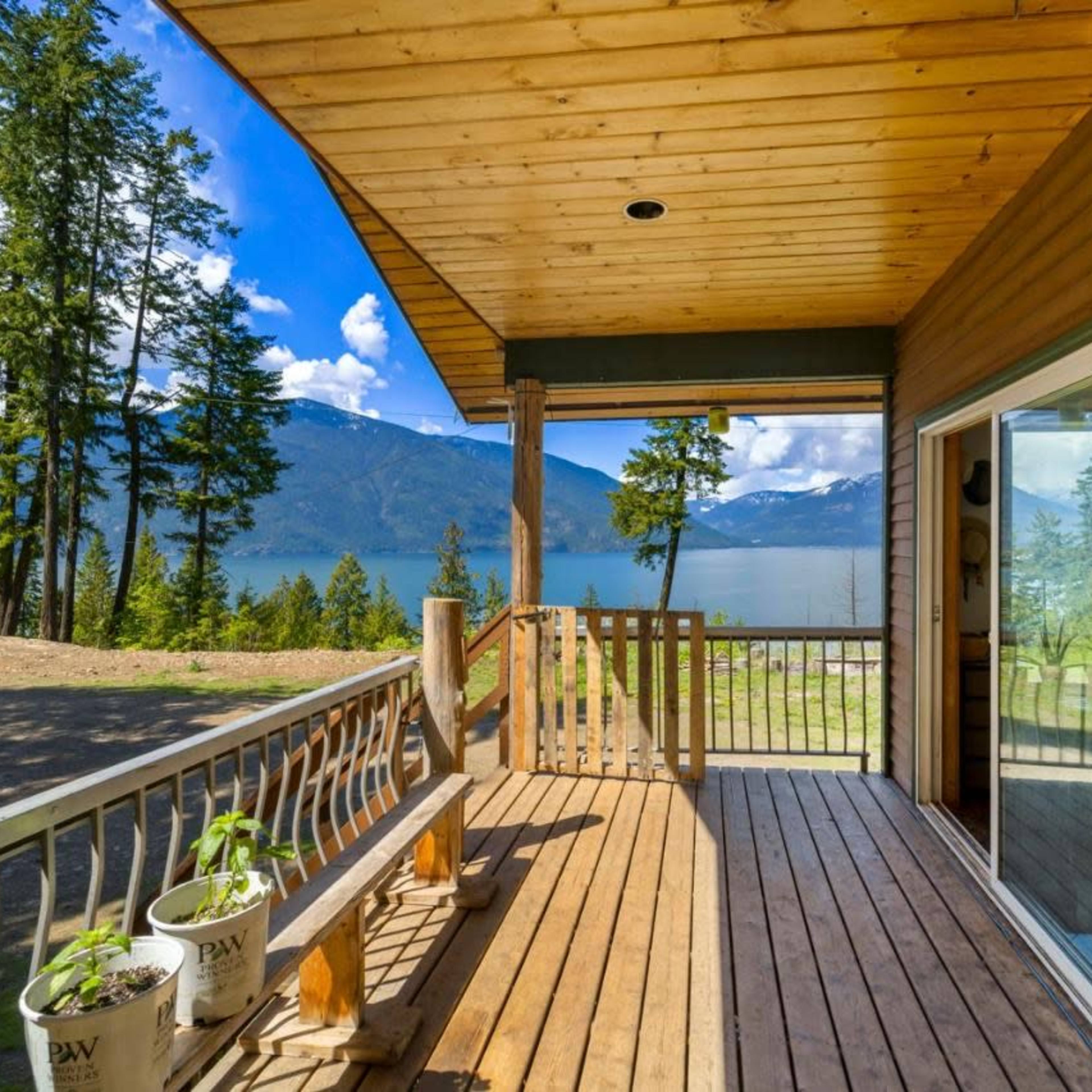 A wooden deck overlooking a lake, surrounded by tall trees and distant mountains under a partly cloudy sky.