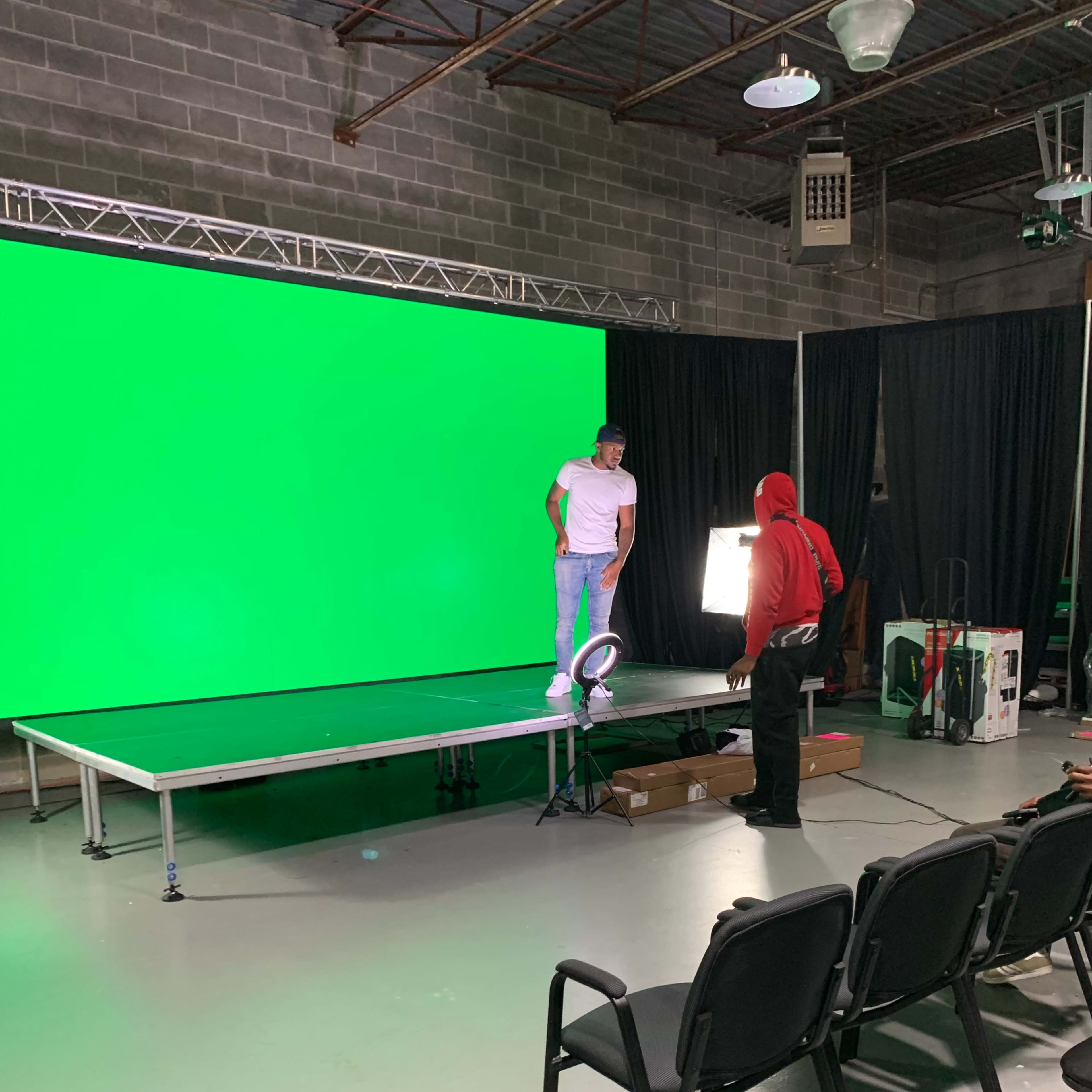A person in a white shirt stands on a platform in front of a large green screen while another person in a red hoodie adjusts lighting equipment nearby.