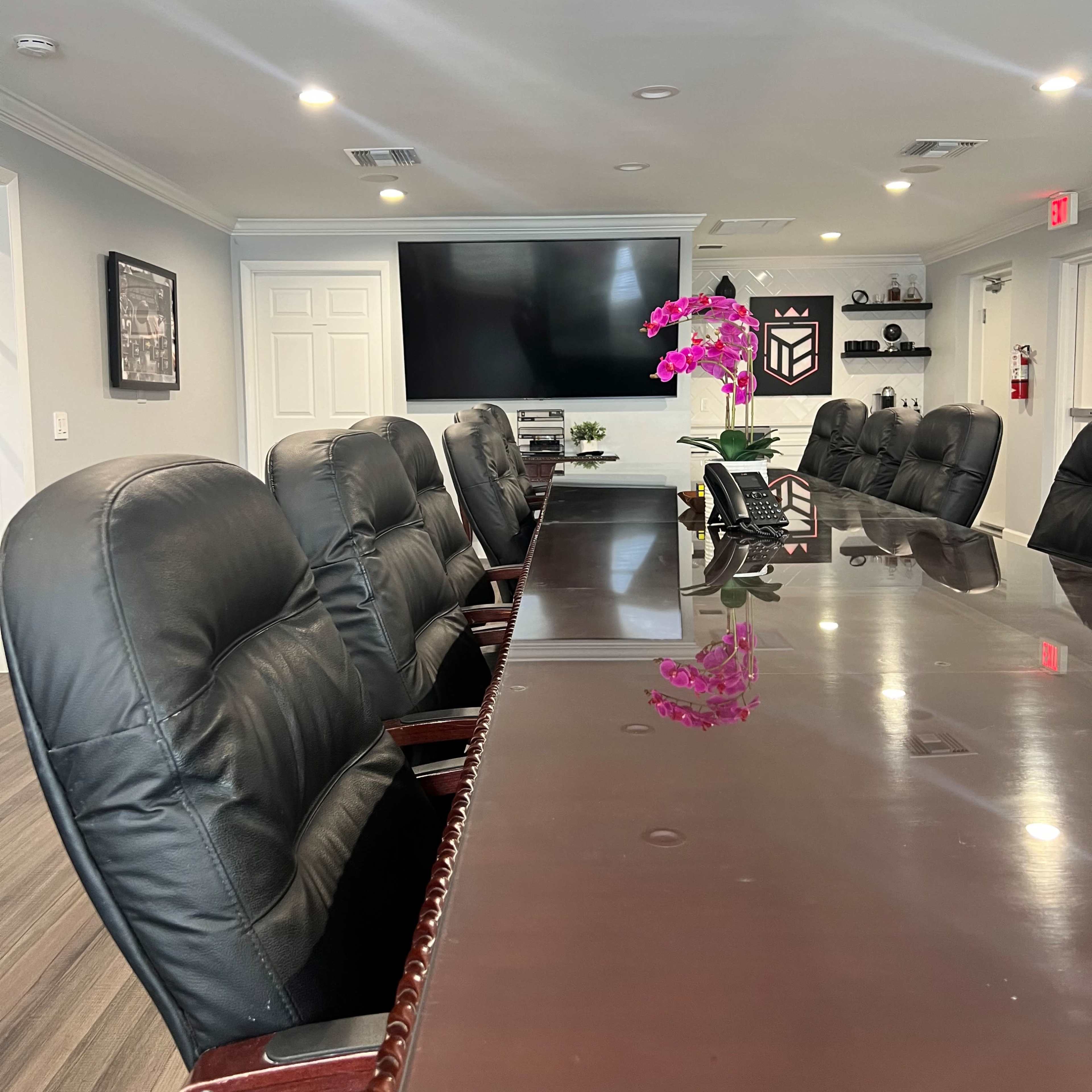 A conference room equipped with a long table, black office chairs, and a large wall-mounted screen at one end.