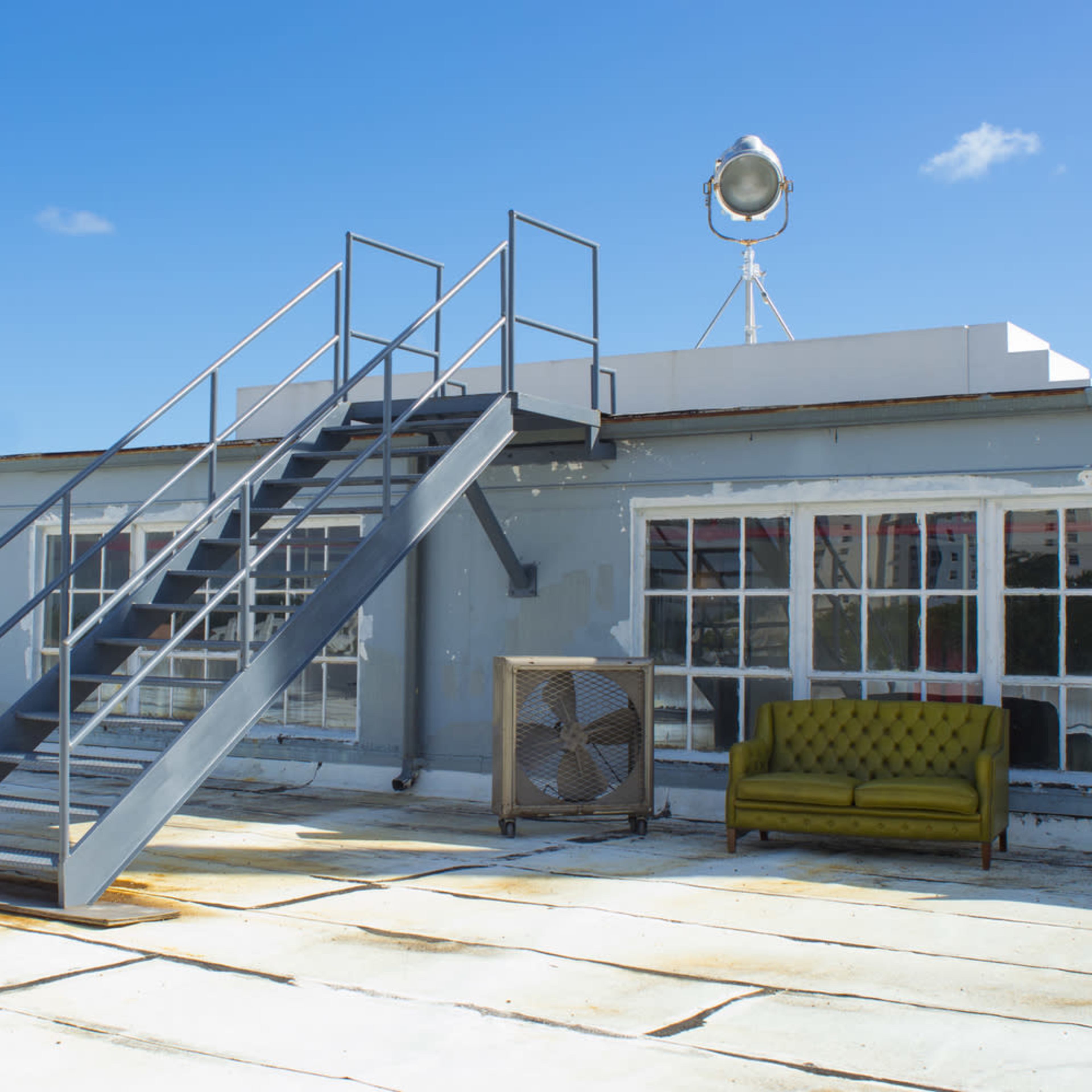 A metal staircase leads up to a rooftop area featuring a large spotlight, with a green sofa and an air conditioning unit nearby.