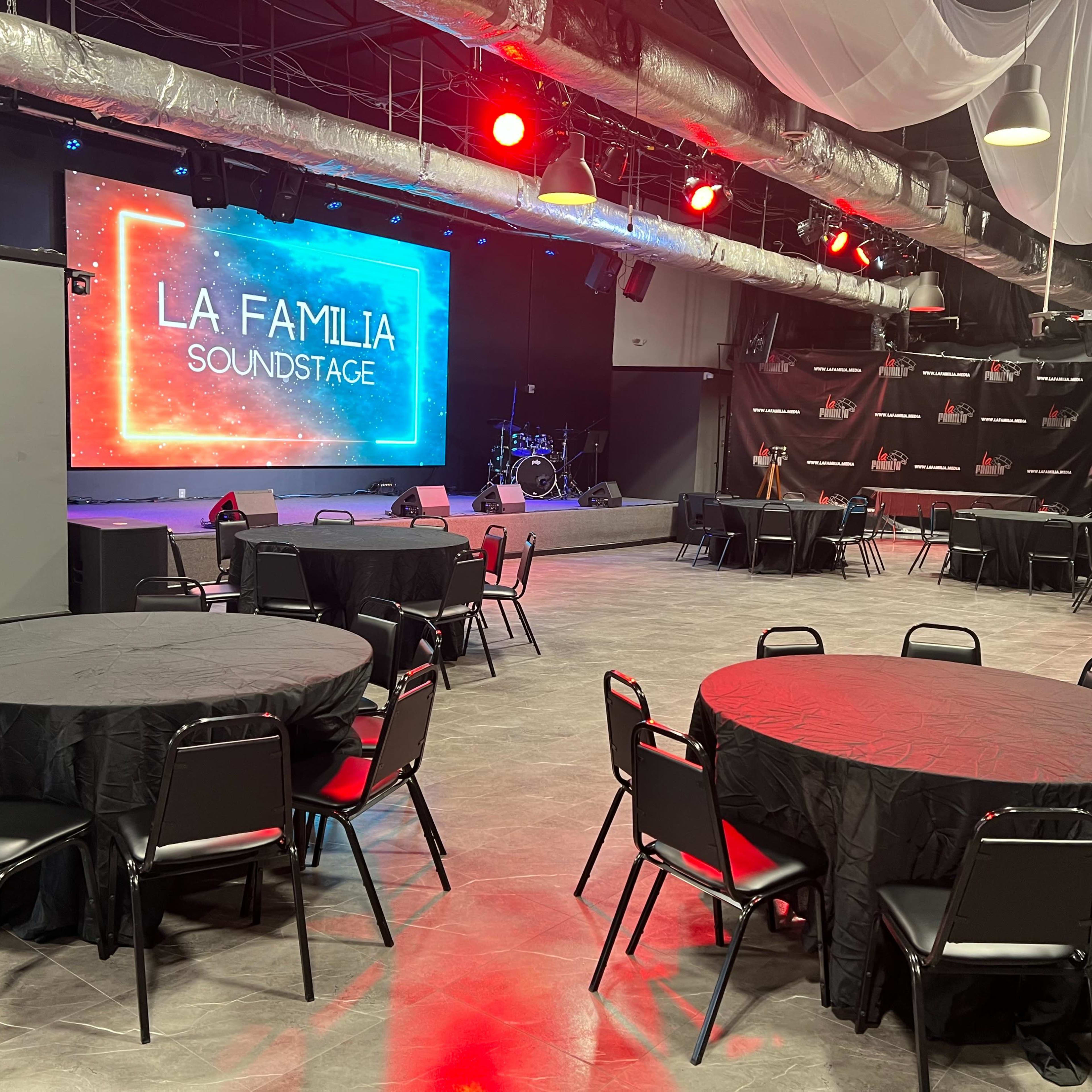 A spacious event venue featuring round black tables and chairs, with a stage in the background displaying a colorful sign that reads "LA FAMILIA SOUNDSTAGE."