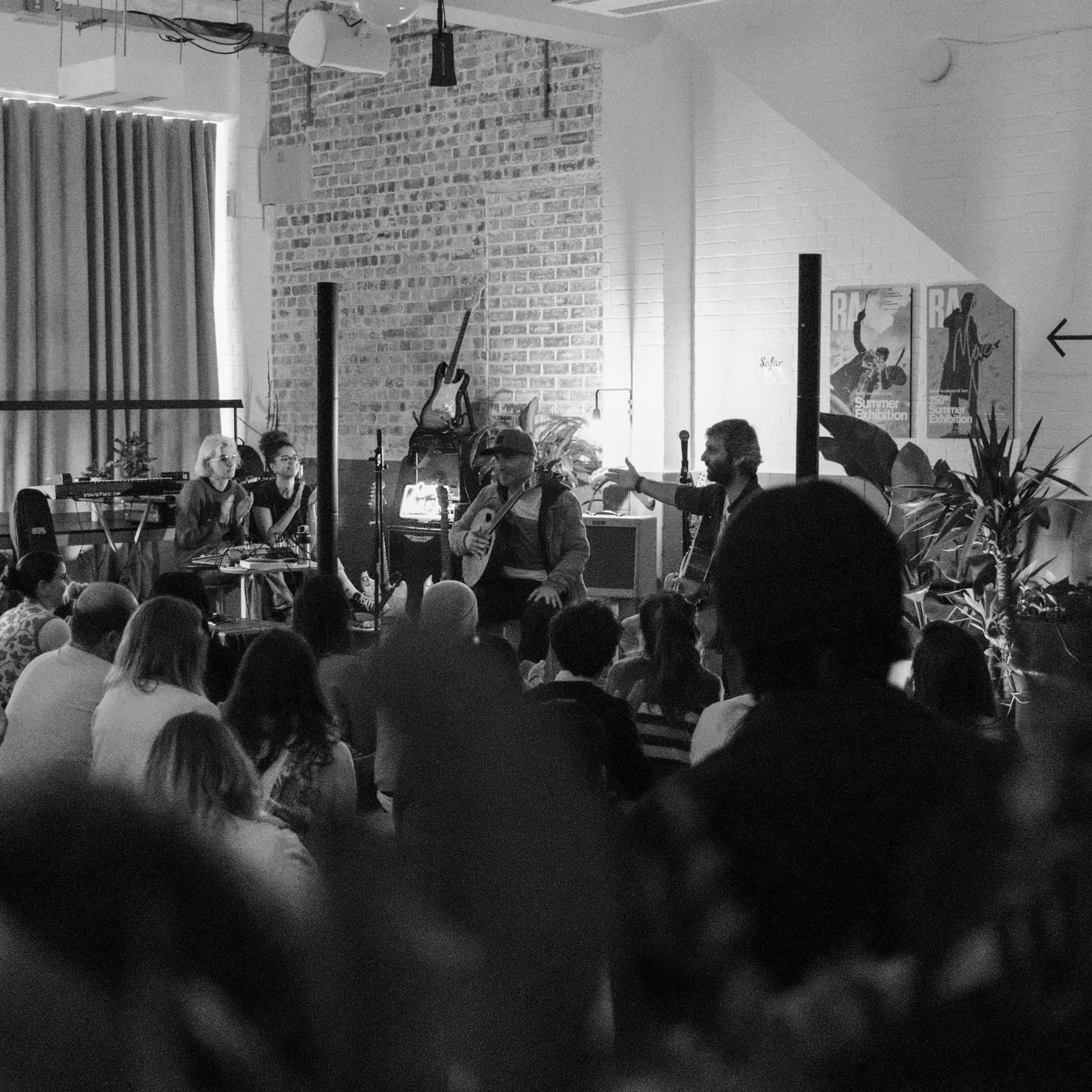 A group of musicians performs for an audience seated on the floor in a dimly lit, indoor space decorated with plants and artwork.