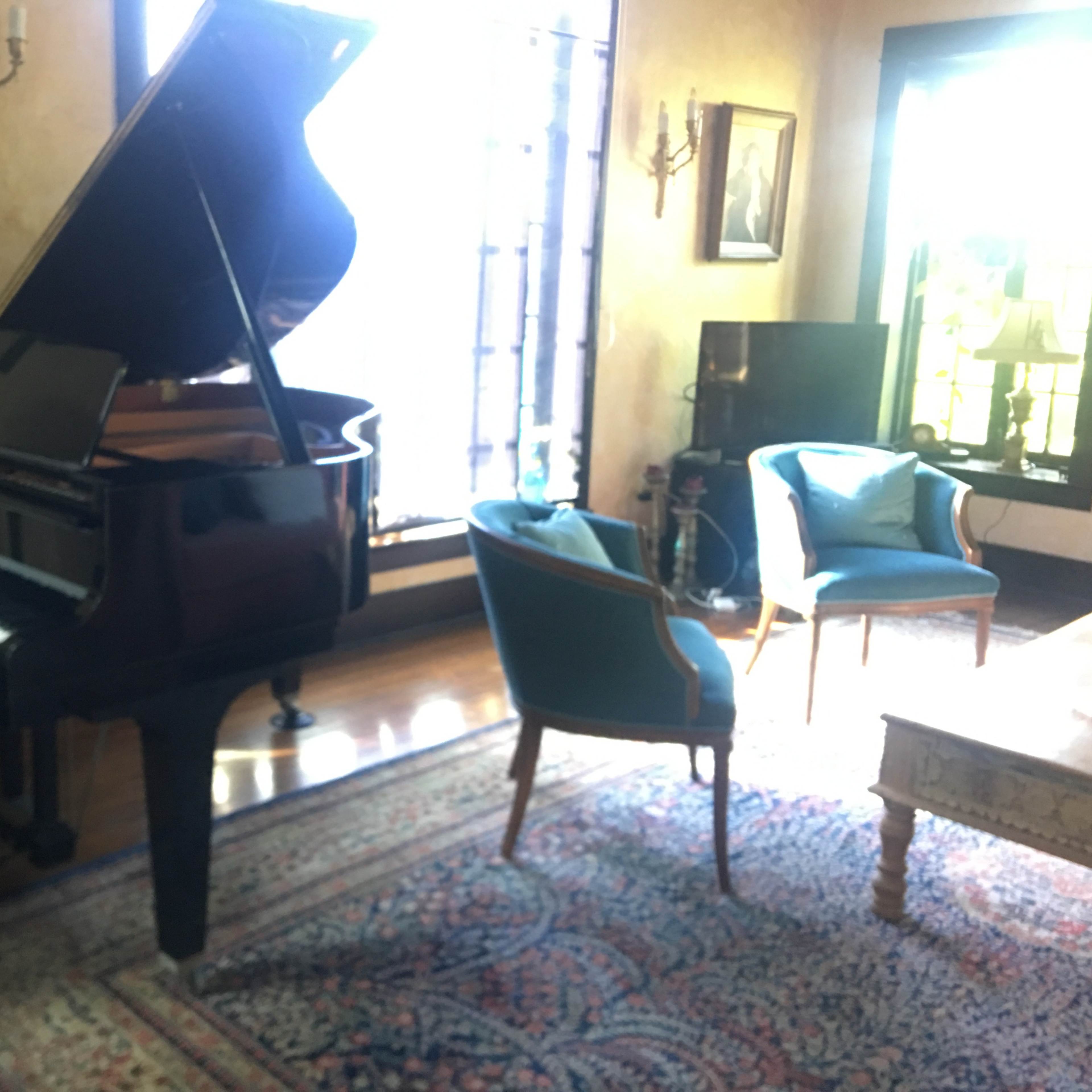 A grand piano stands next to a pair of upholstered chairs in a well-lit living room with large windows and a patterned rug.