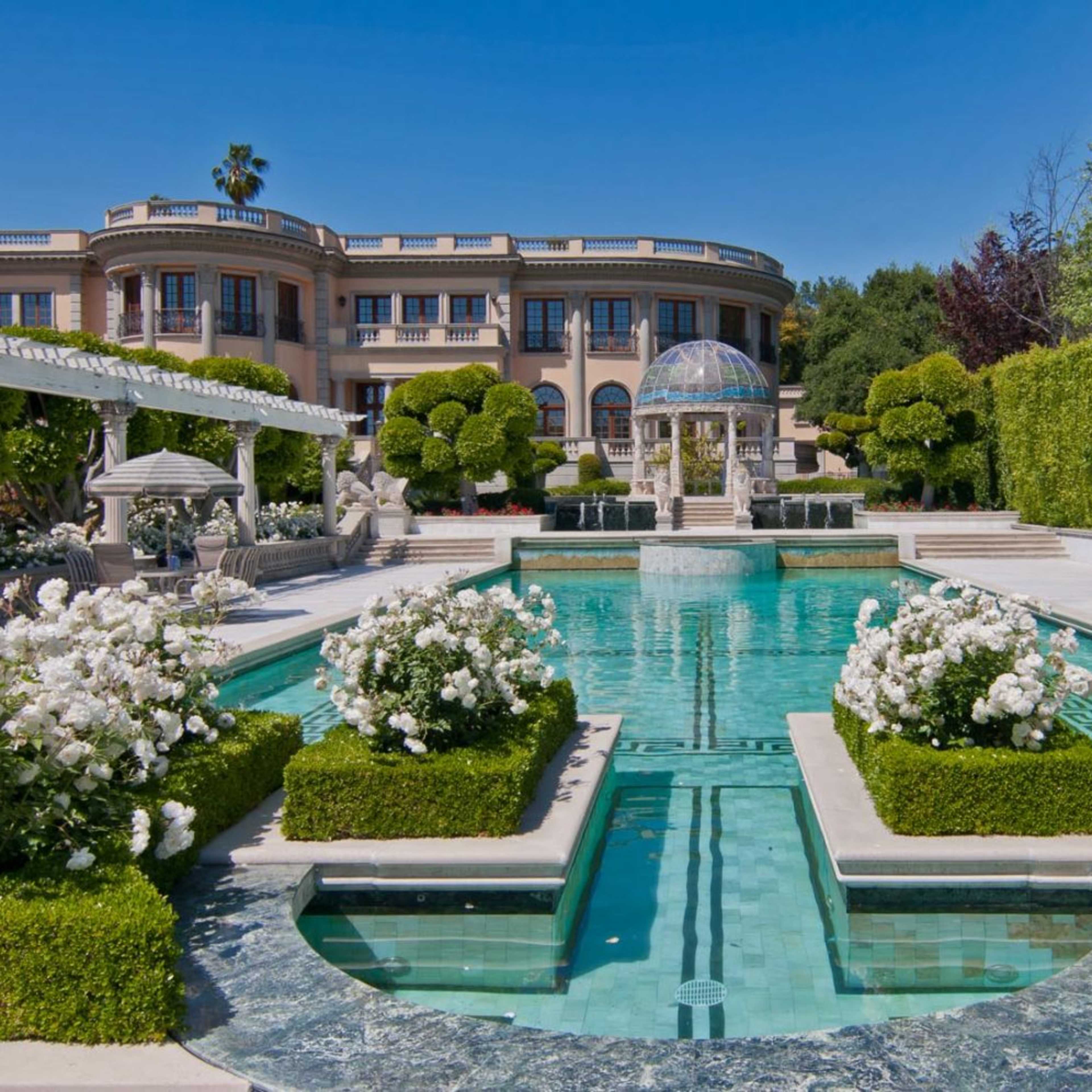 A large swimming pool is surrounded by manicured gardens and a lavish mansion in the background.