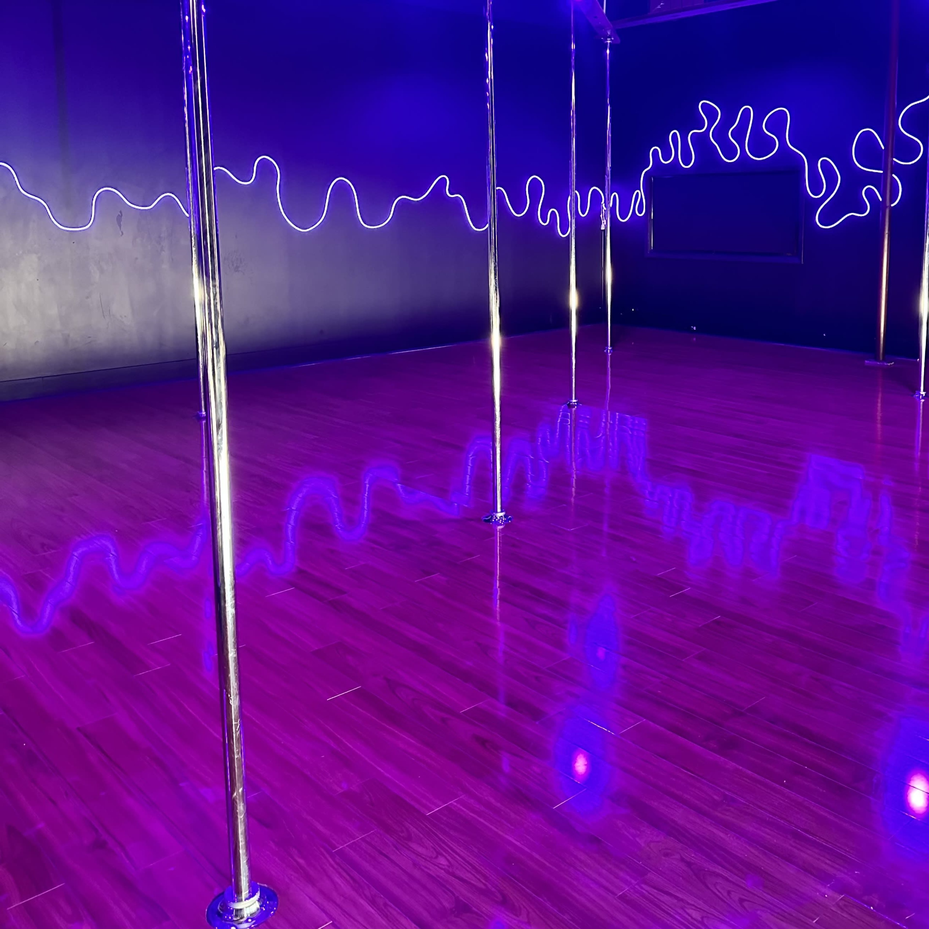 A dance studio with polished wooden flooring and several metallic poles, illuminated by purple neon lights along the walls.