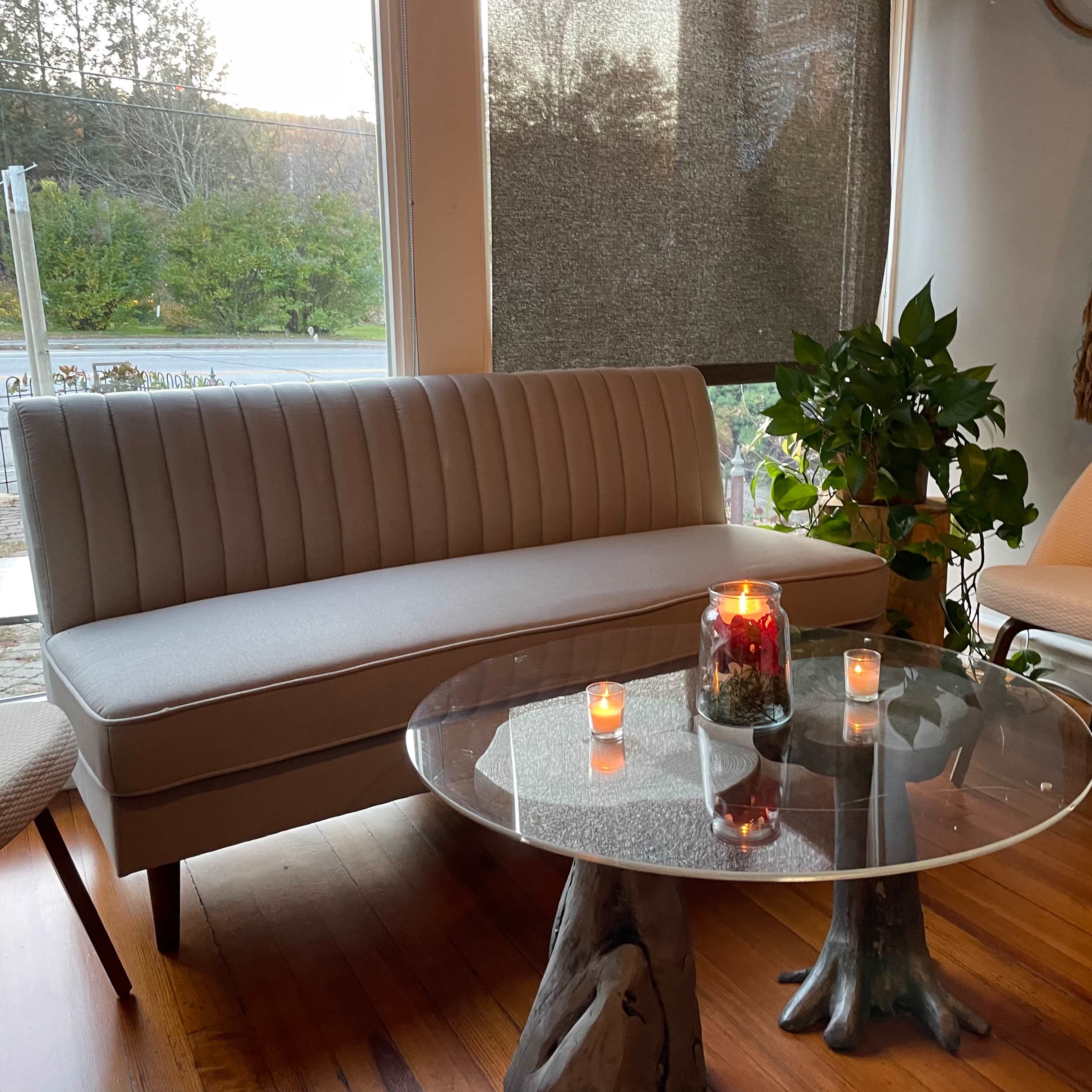 A modern seating area features a light sofa, a glass table with candles, and potted plants beside large windows.