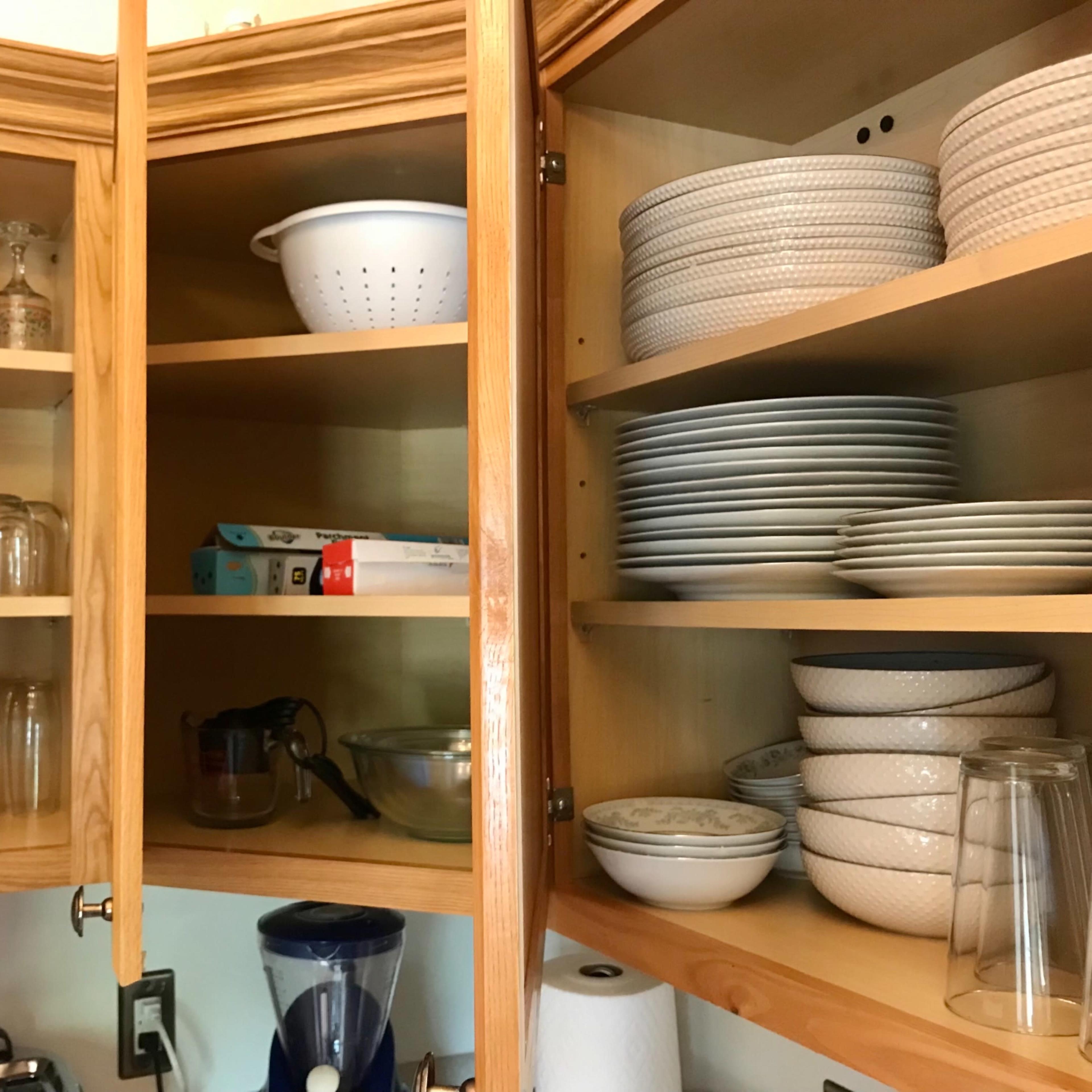 A wooden kitchen cabinet with glassware, a colander, and plates neatly arranged on multiple shelves.