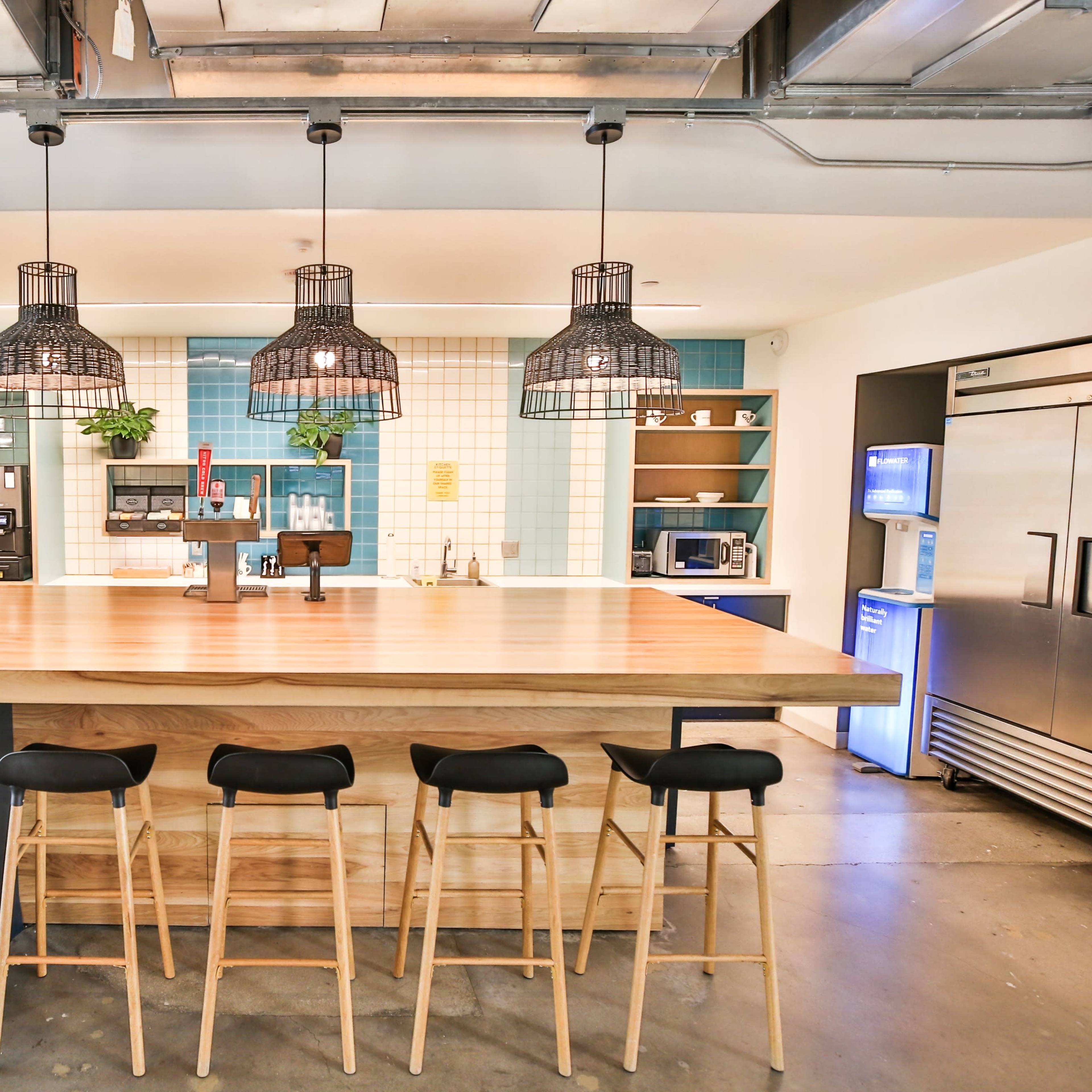 A modern kitchen features a large wooden island with four stools, pendant lights hanging above, and shelves displaying plants and appliances in the background.