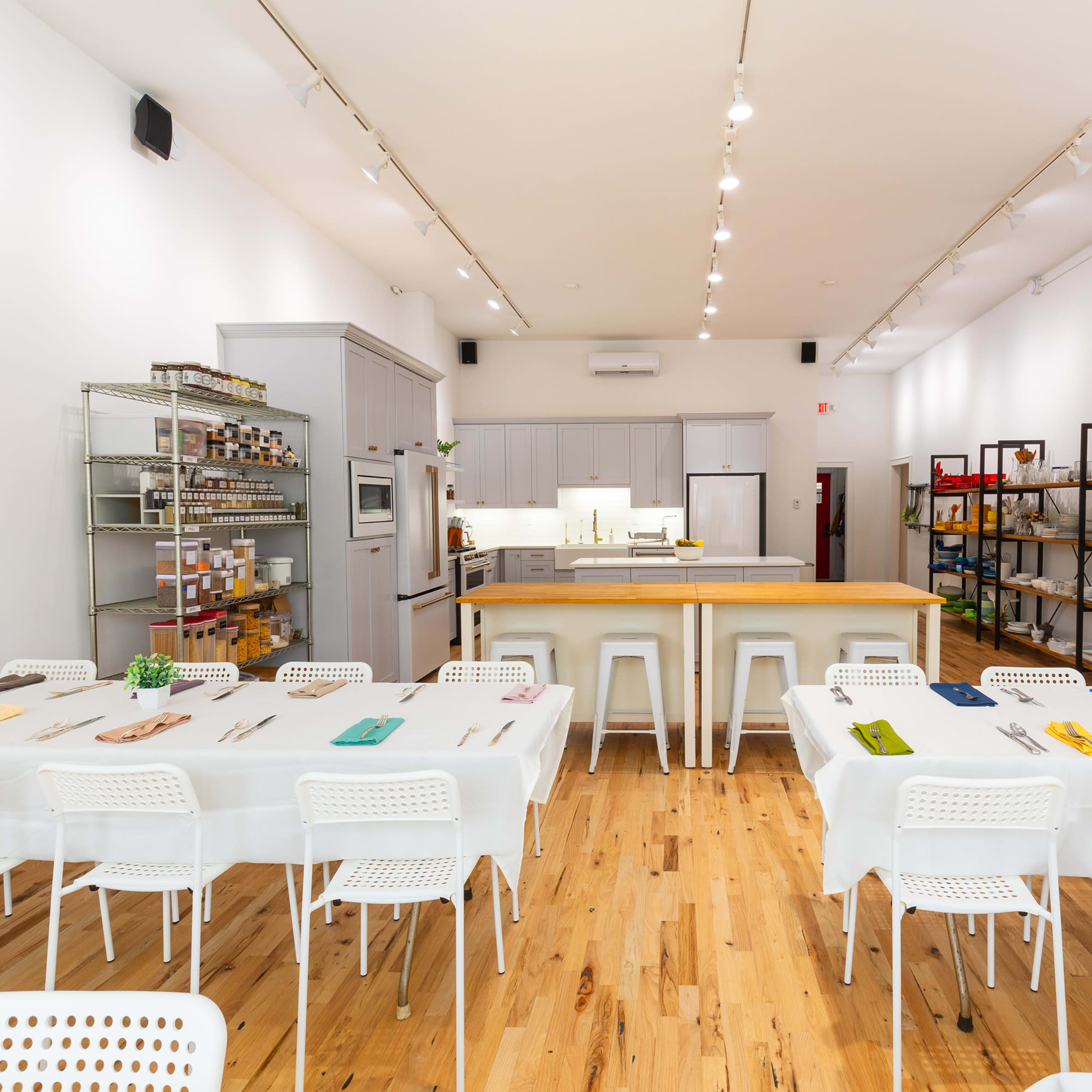 A bright, modern cooking classroom with a central kitchen island surrounded by neatly arranged tables and chairs.