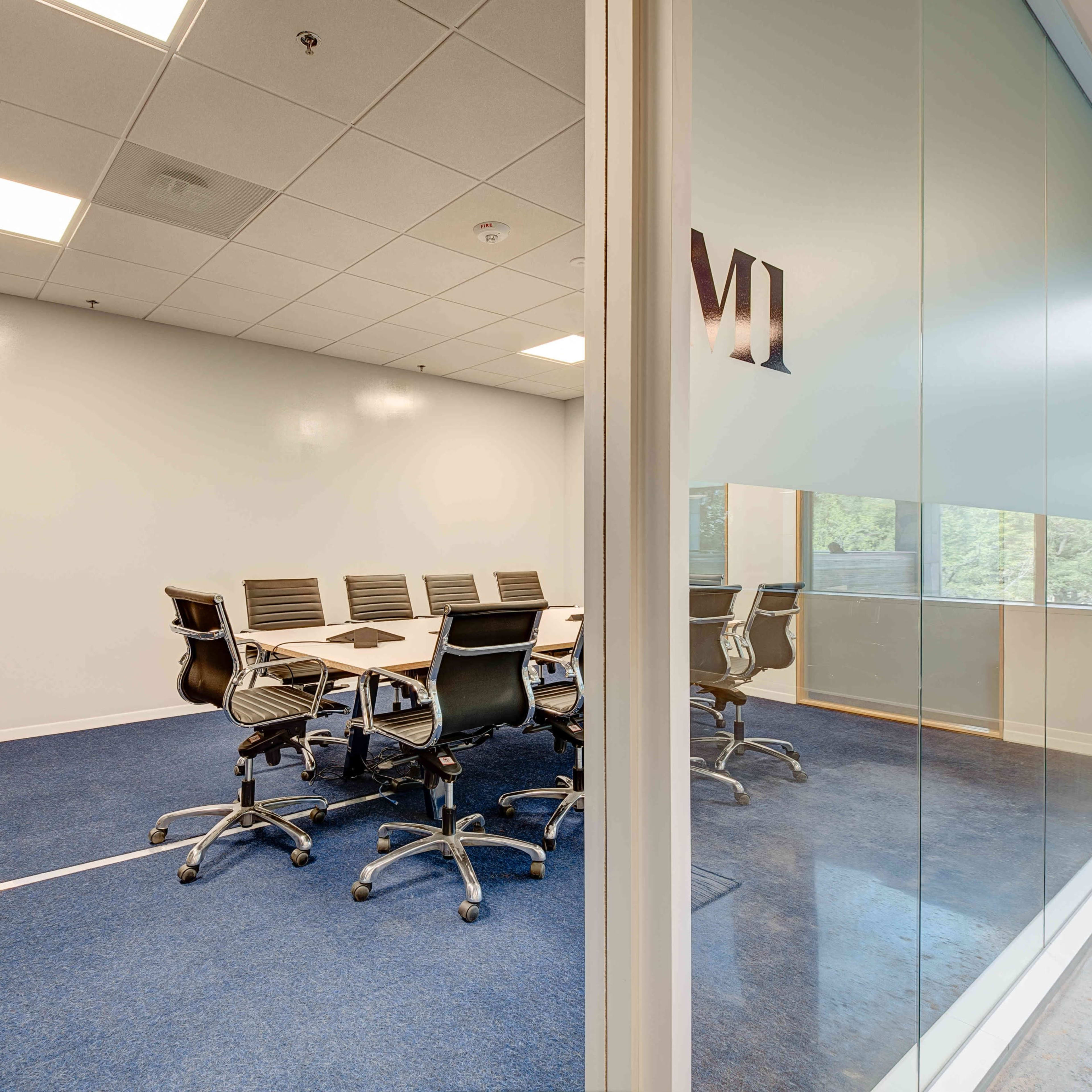 A modern conference room with a glass wall features a large table surrounded by black chairs and a television on one side.