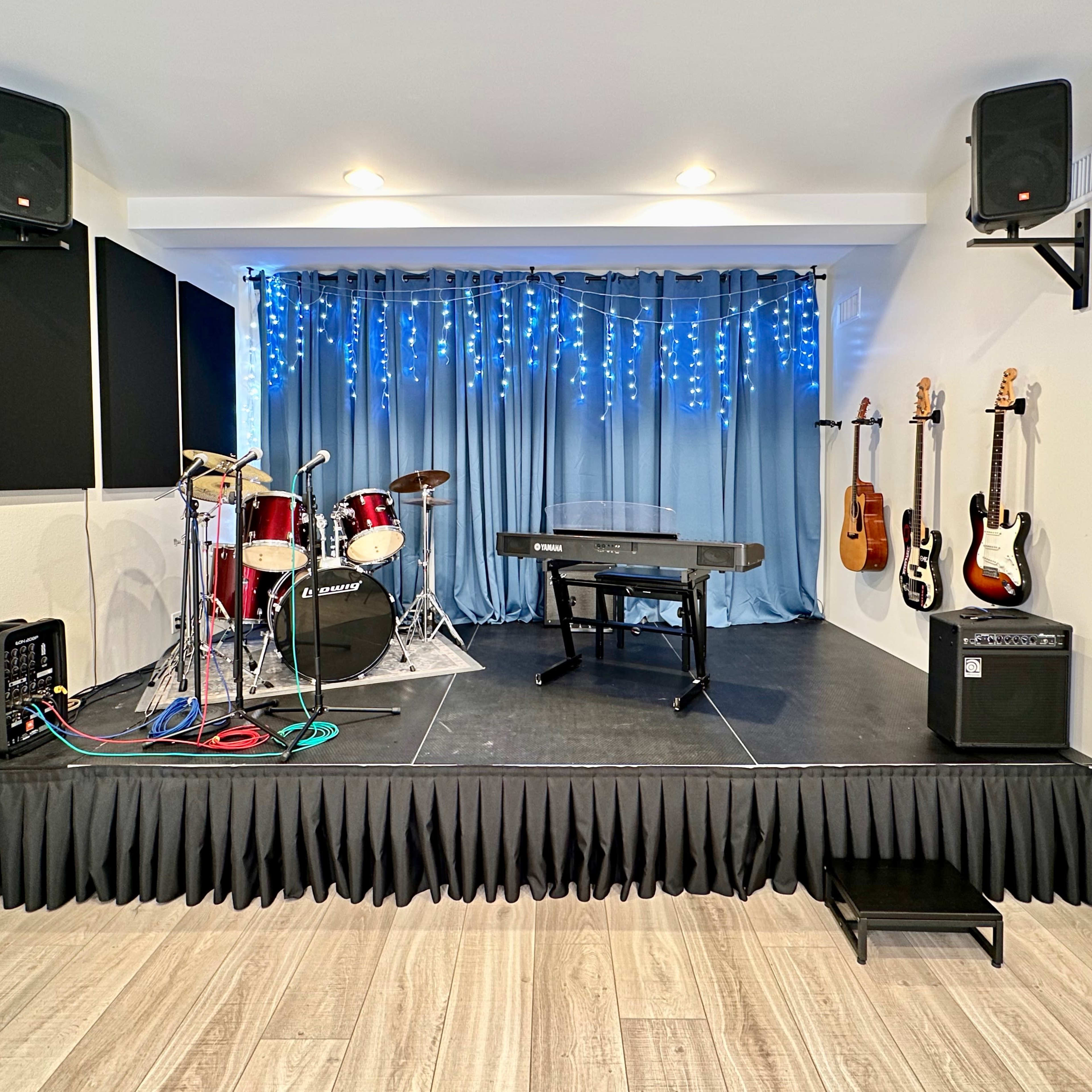 A small stage setup featuring a drum kit, a keyboard, and several electric guitars mounted on the wall, with blue curtains and string lights in the background.