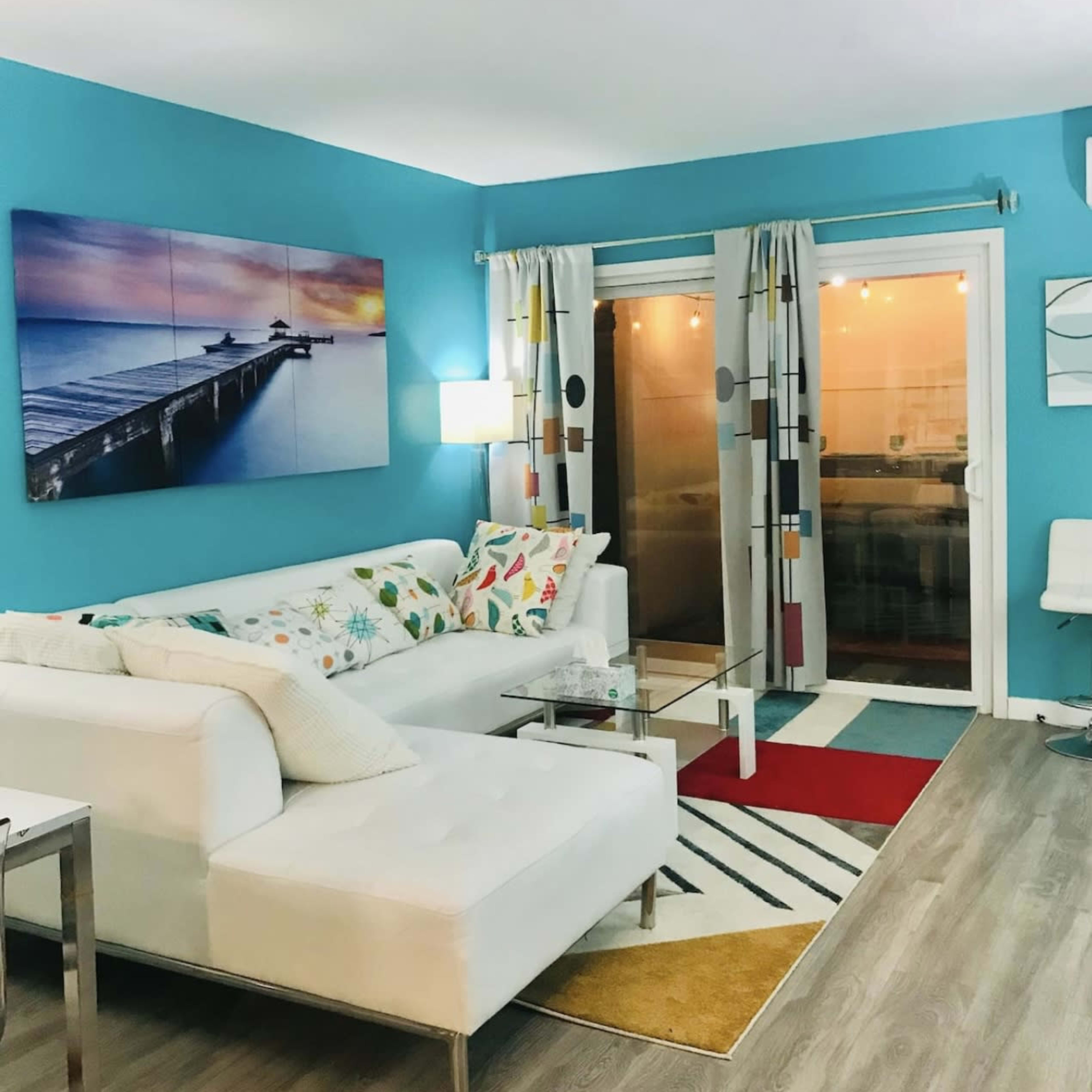 A brightly colored living room with a white sectional sofa, a glass coffee table, and a large piece of wall art featuring a pier at sunset.