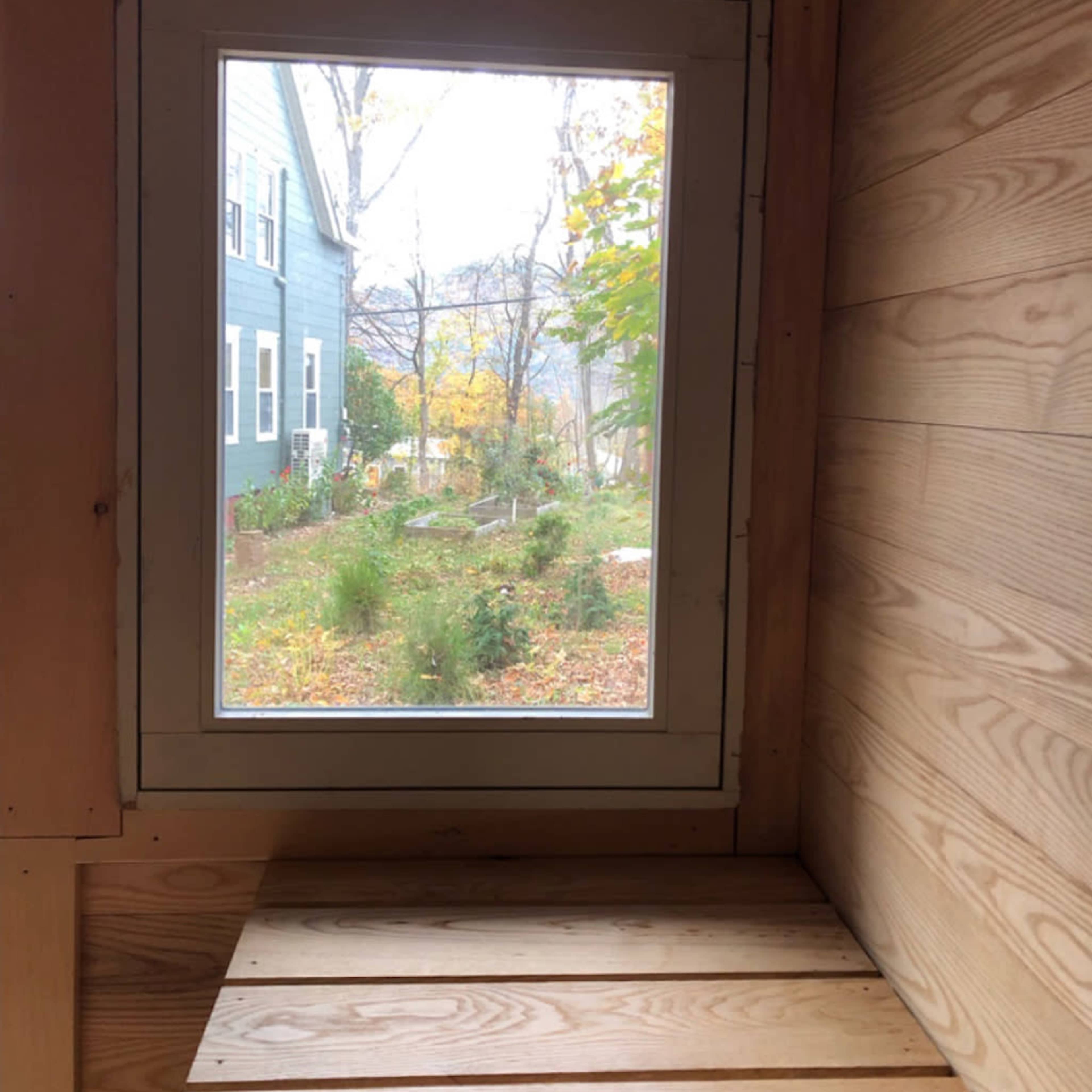 A wooden interior with a window framing a view of an outdoor garden and nearby houses.