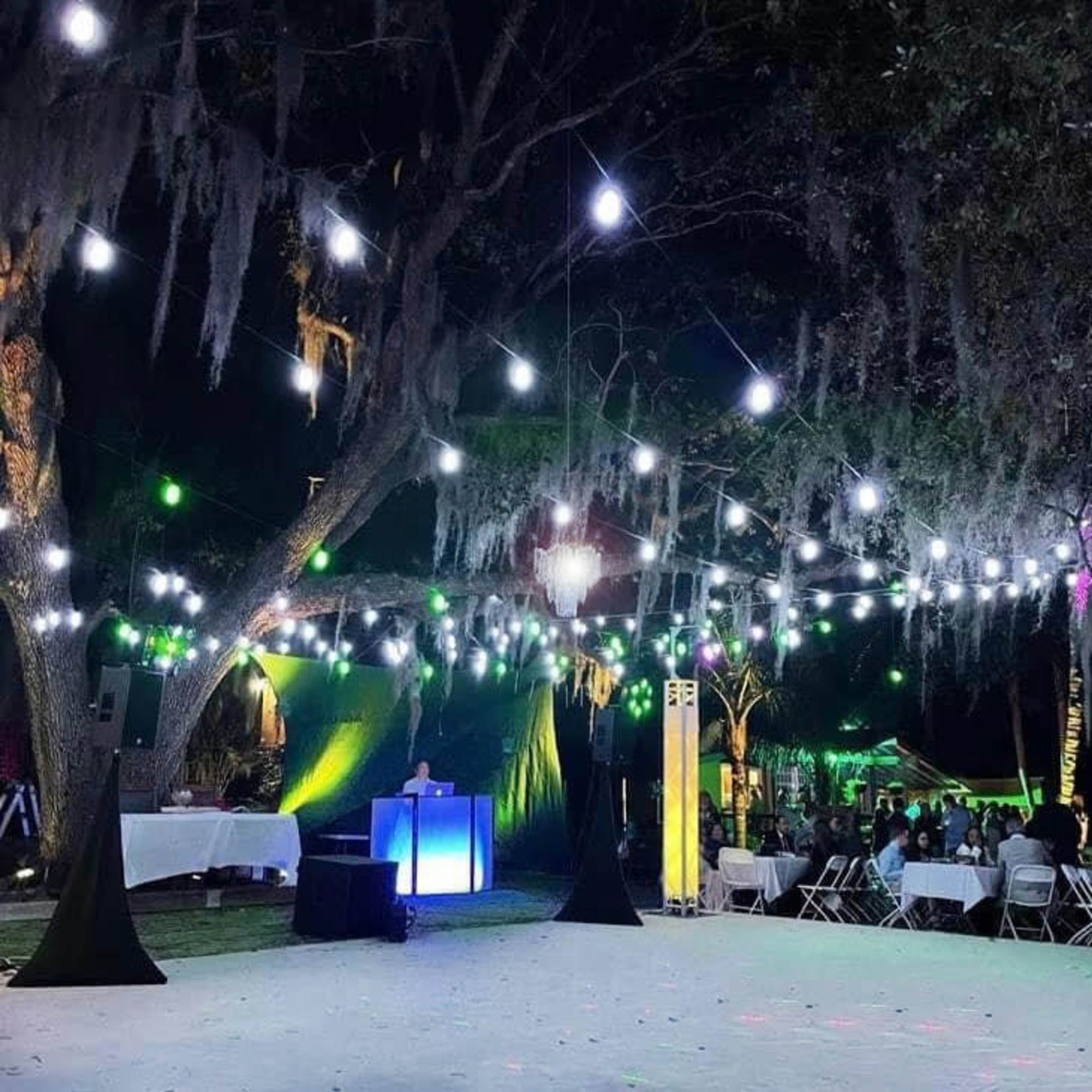 A decorated outdoor event space features tables and chairs under string lights and illuminated trees at night.