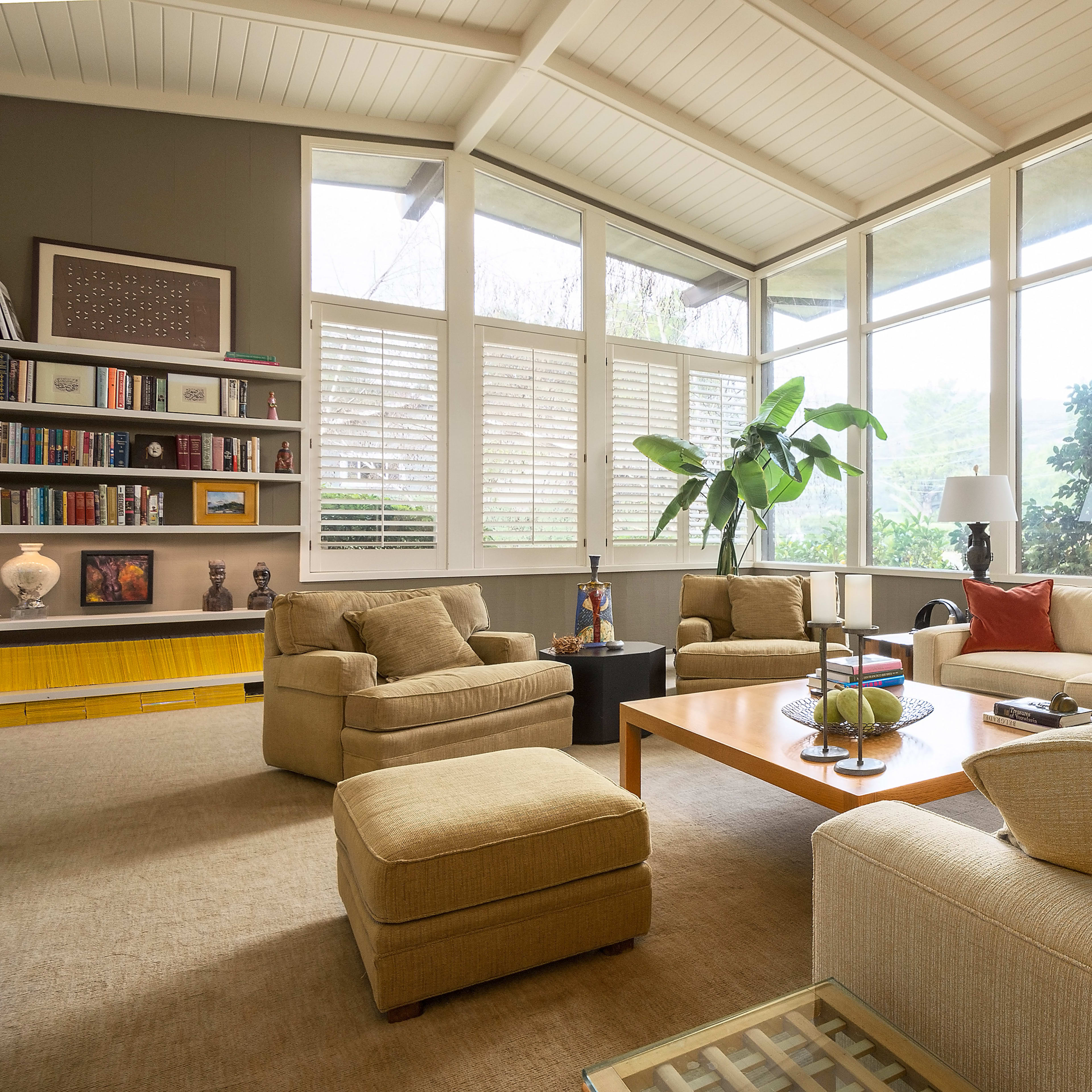 A bright, modern living room with large windows, comfortable beige sofas, and a coffee table surrounded by armchairs.