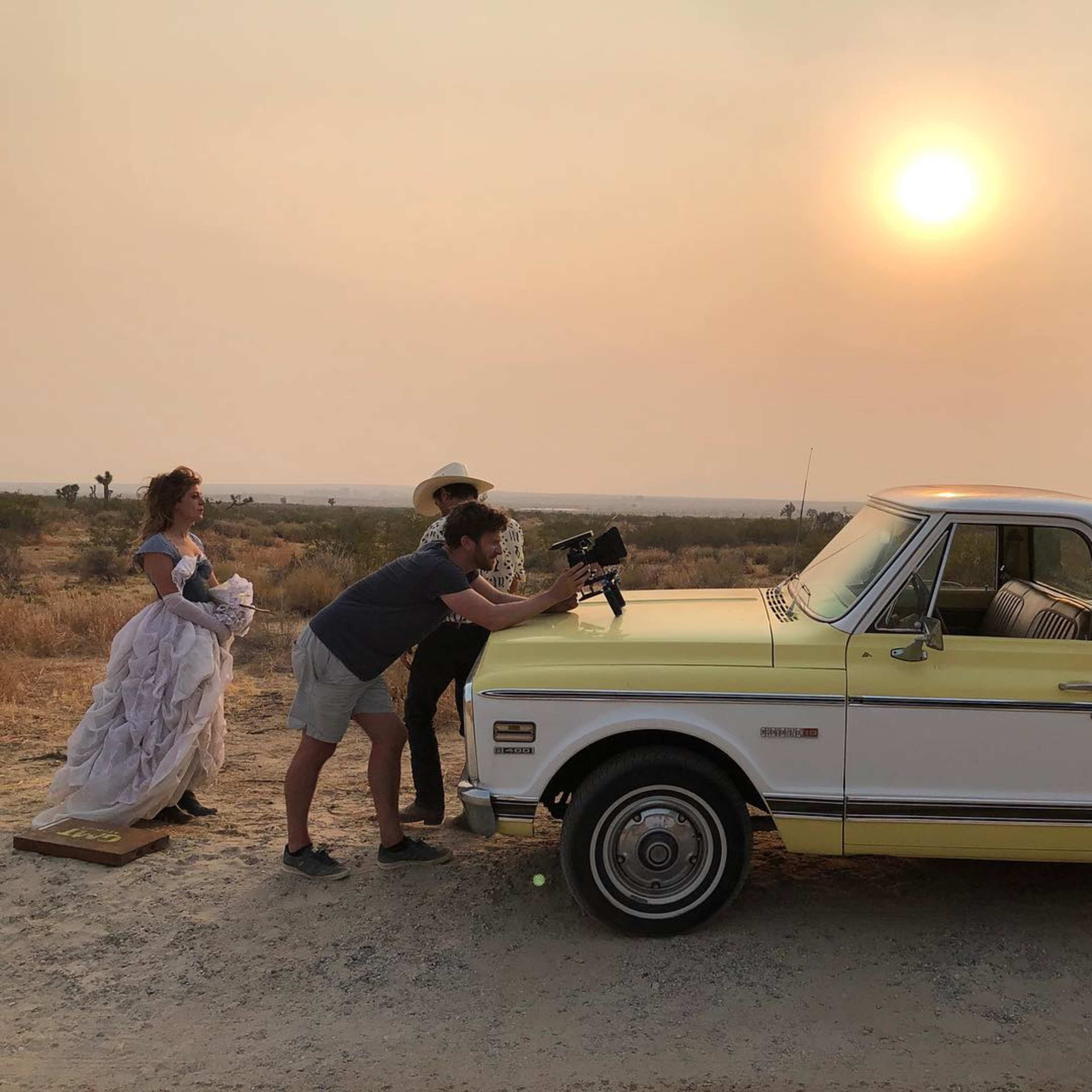 Three people set up a camera in front of a vintage yellow and white pickup truck on a dusty road under a hazy sky with a setting sun.
