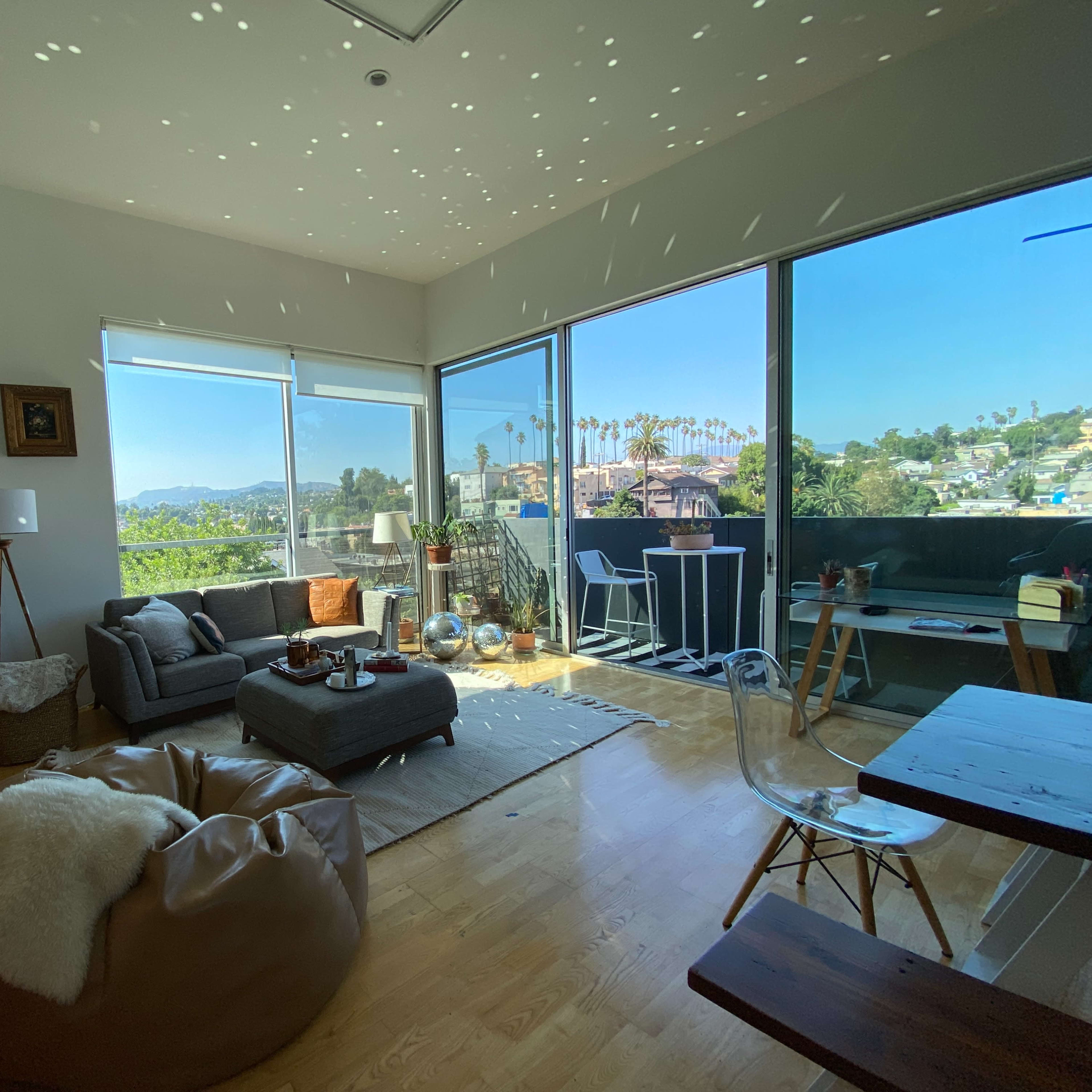 A modern living room features a gray sofa, a large window with a view of palm trees, and light spots dancing on the ceiling.