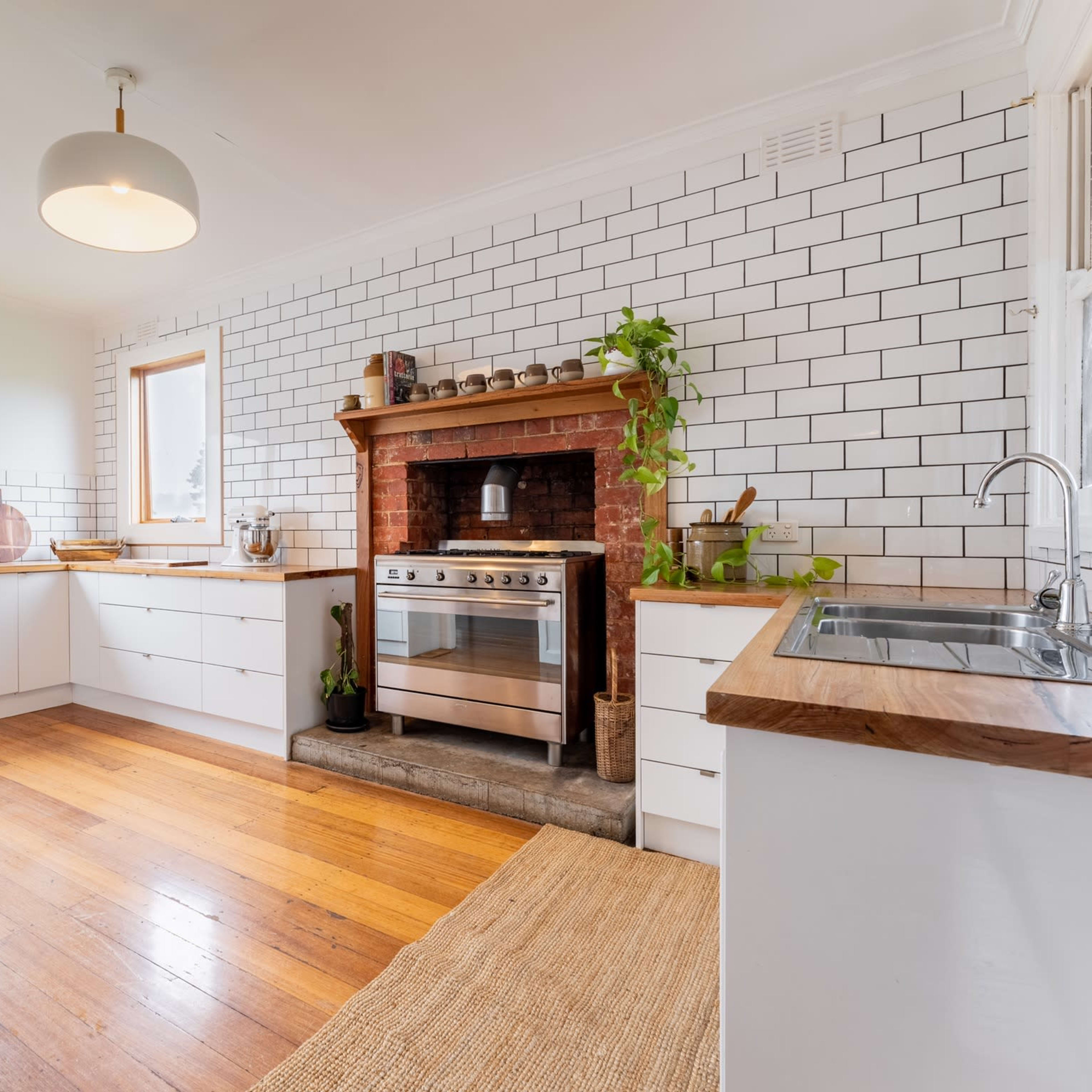 A modern kitchen features white cabinetry, a brick fireplace with an oven, and wooden floors, illuminated by natural light through large windows.