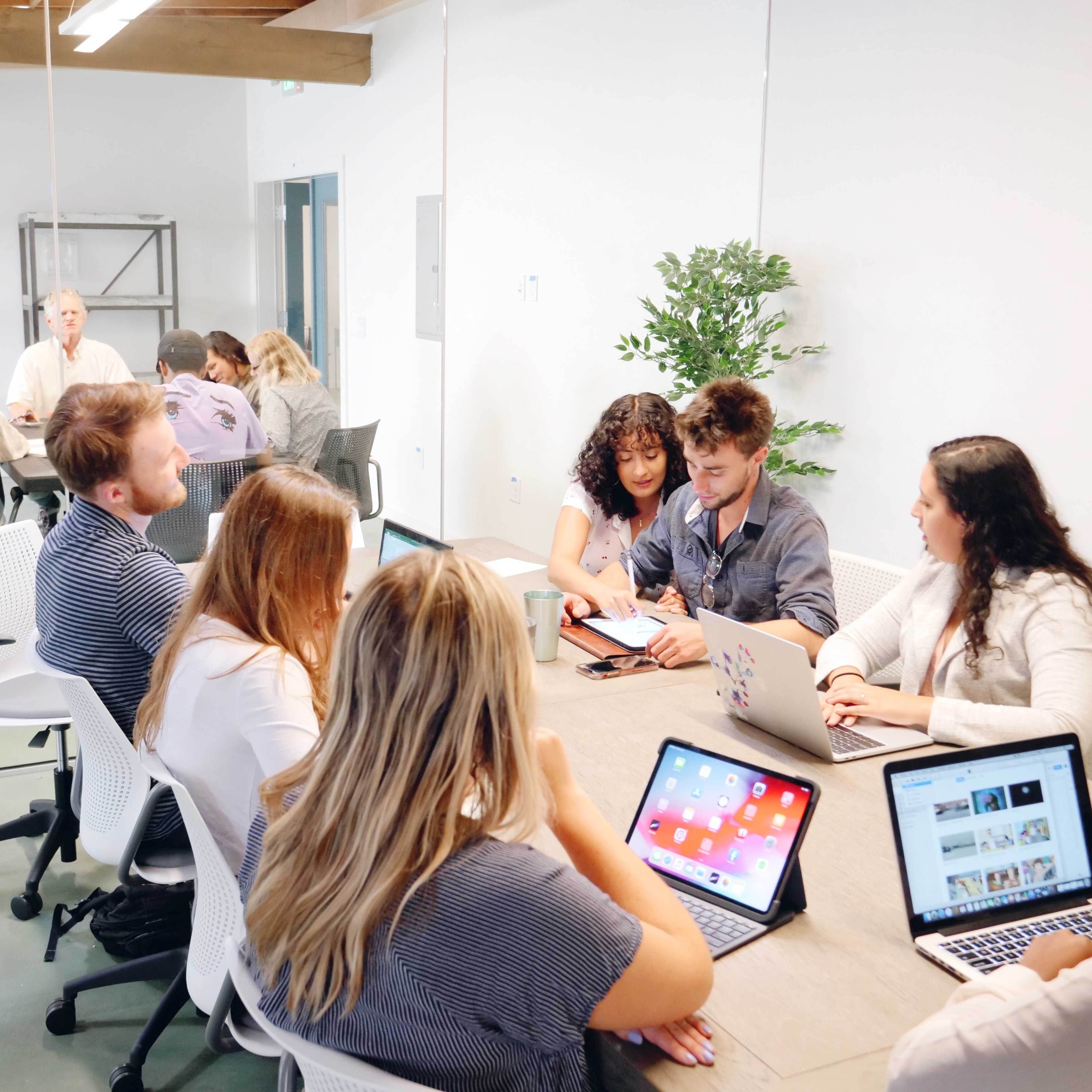 A group of people is seated around a large table in a conference room, engaging in discussions and working on laptops.