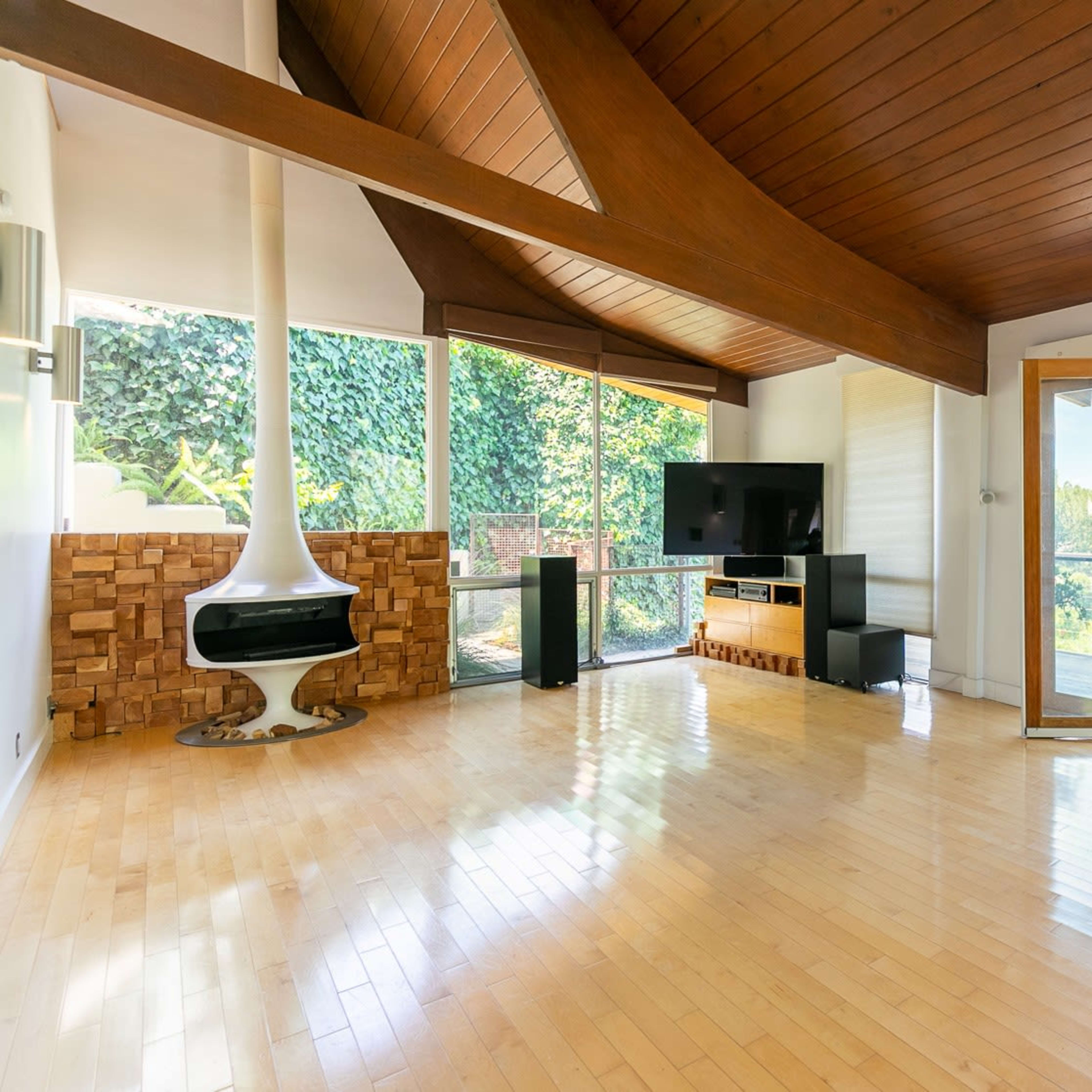 A modern living room with large windows, wooden beams, and a white fireplace, overlooking a lush outdoor view.
