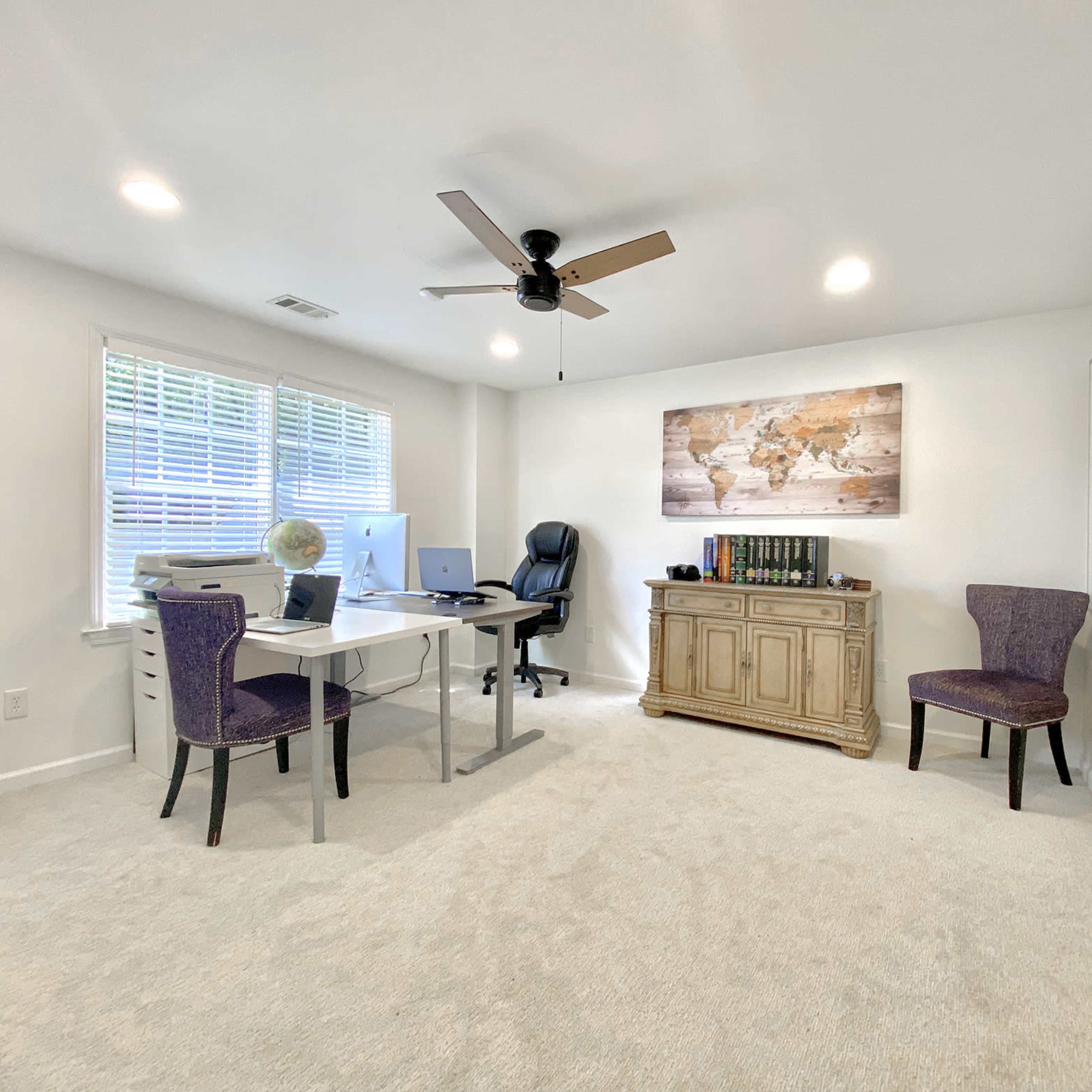 A home office with a desk, an ergonomic chair, two purple chairs, and a wooden cabinet, all situated in a brightly lit room with carpeted flooring.