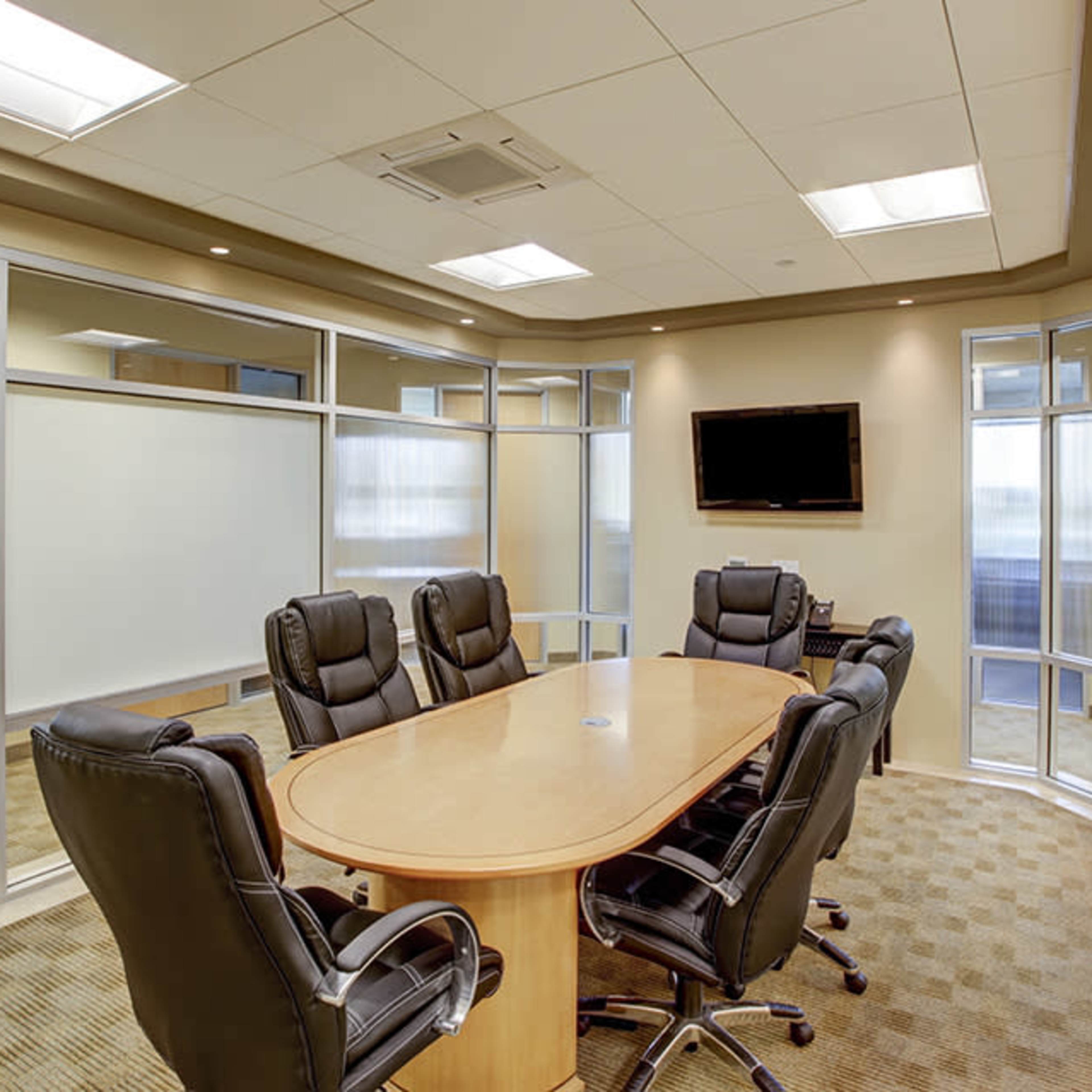 A modern conference room with a long wooden table surrounded by six leather chairs and a wall-mounted television.