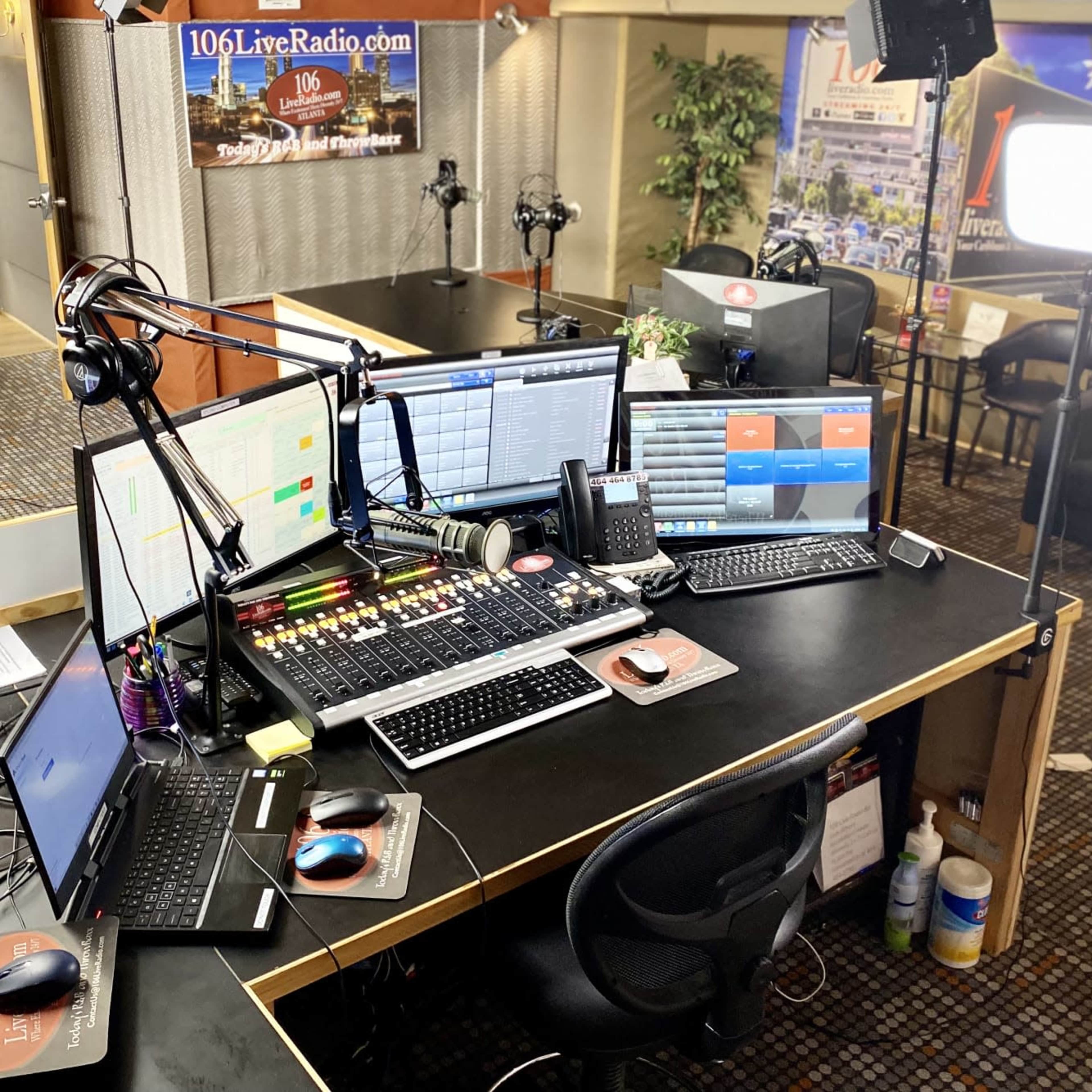 A radio studio with multiple computer monitors, a control board, microphones, and a cozy seating area in the background.