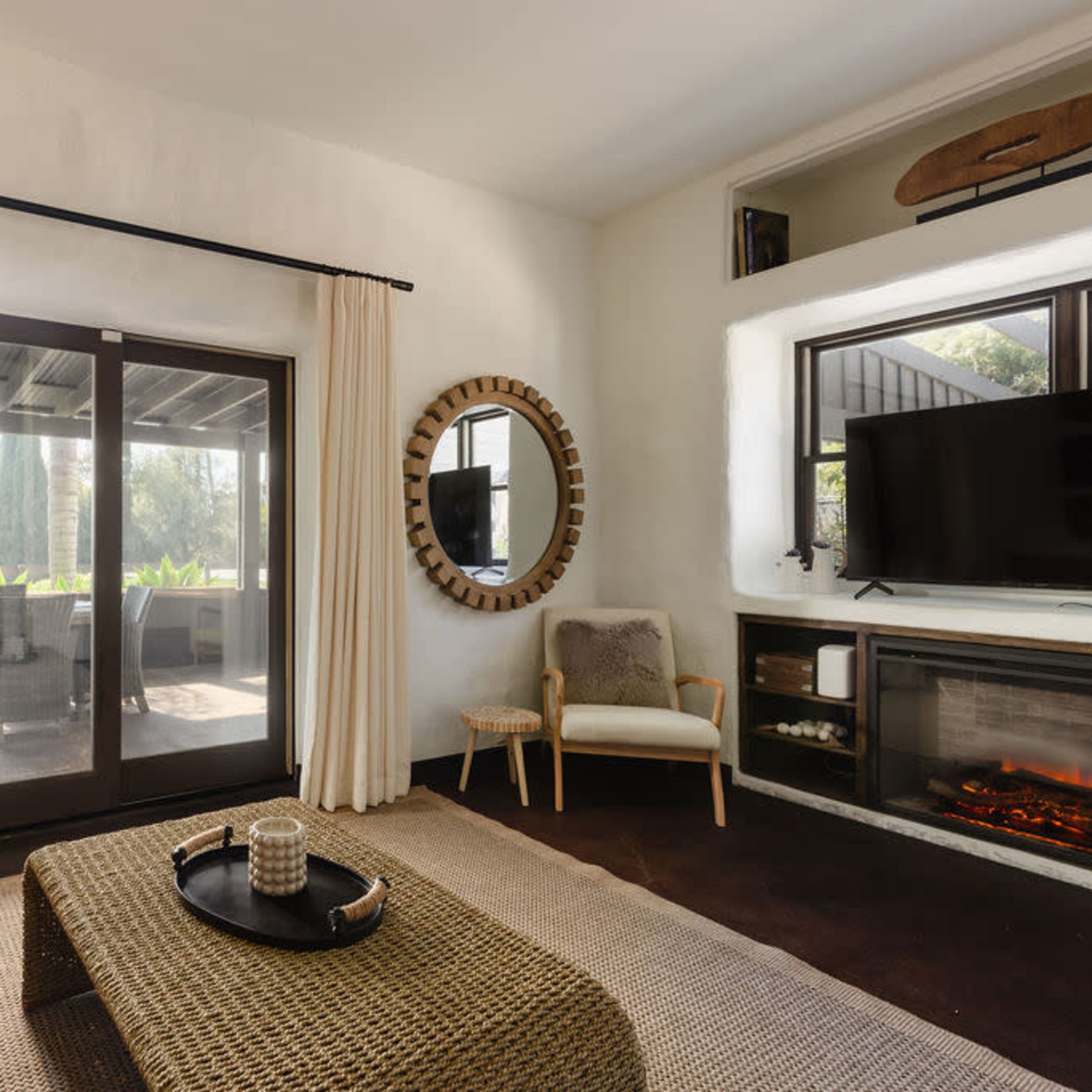 A cozy living room with a leather sofa, a round mirror, a television mounted on the wall, and large glass doors leading to an outdoor patio.