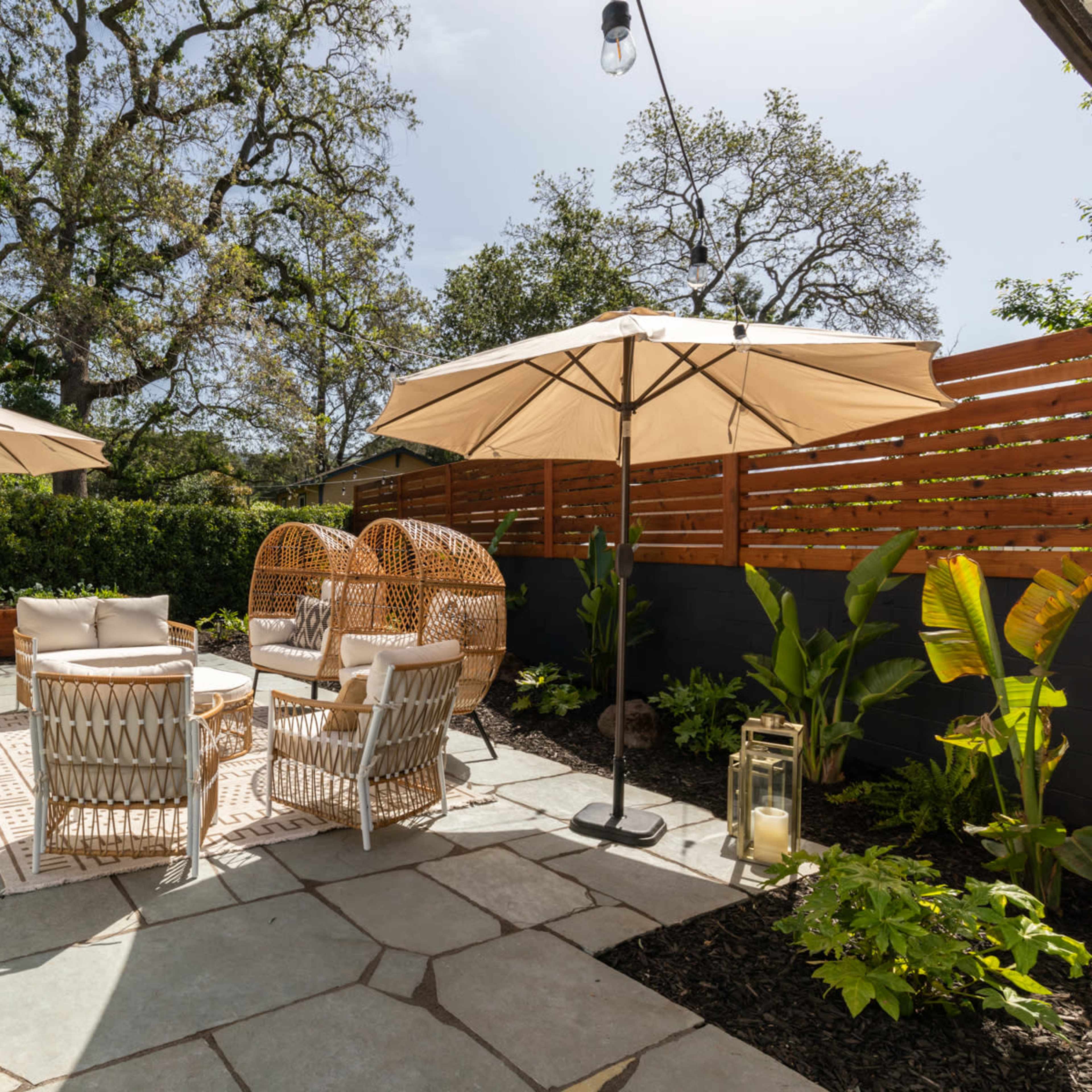 A garden patio featuring four chairs, two decorative umbrellas, and surrounding greenery.