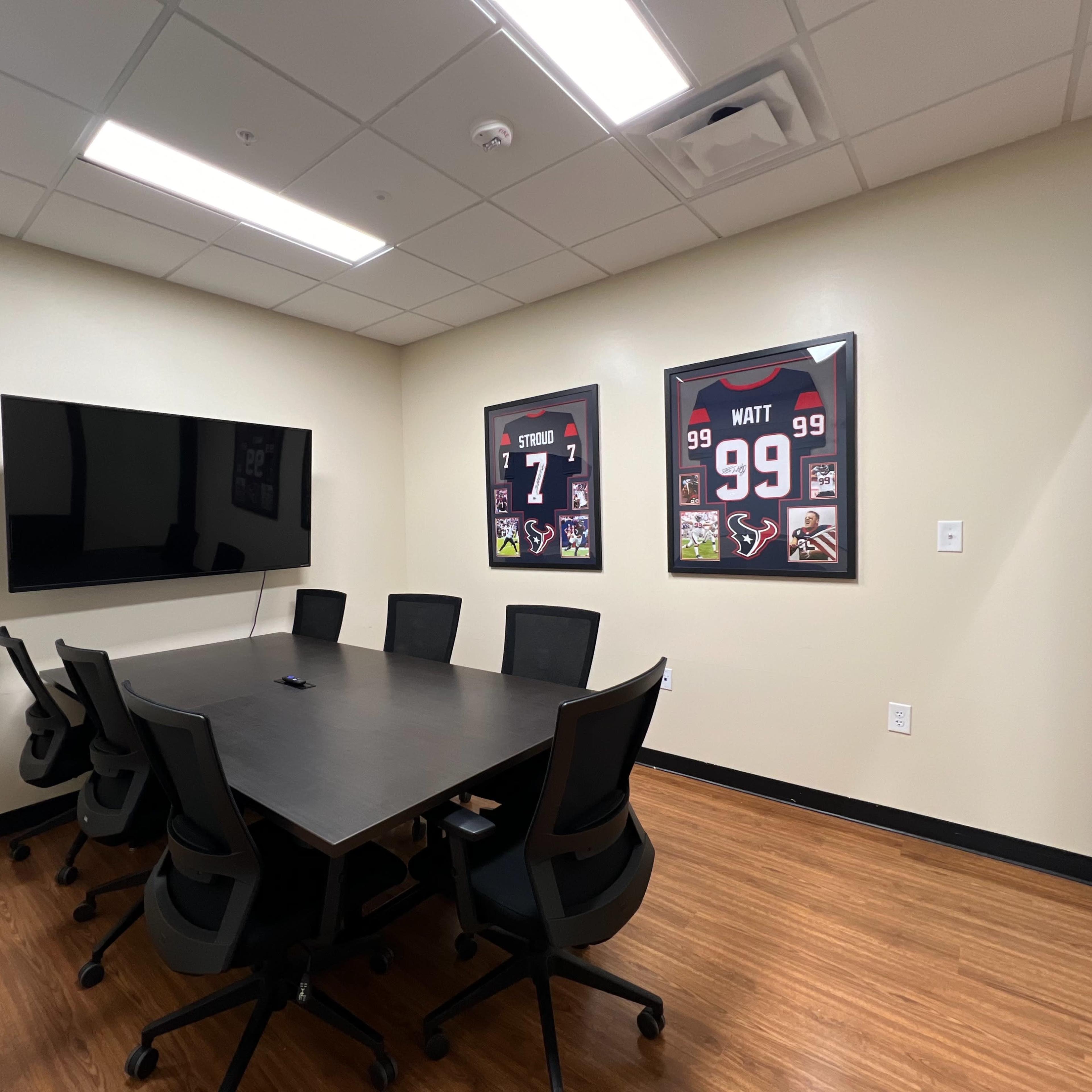 A conference room with a large table surrounded by six chairs, a wall-mounted TV, and two framed sports jerseys displayed on the walls.
