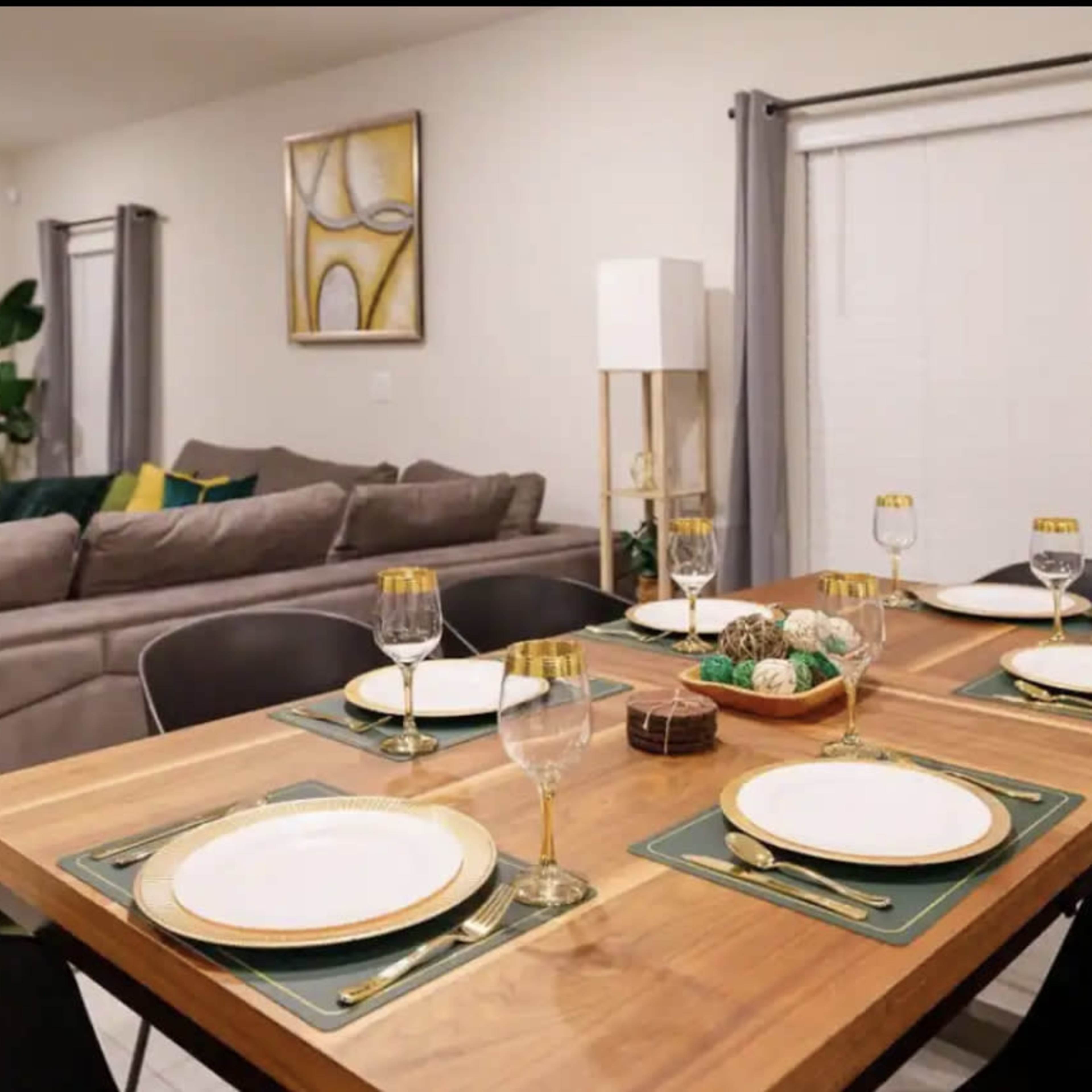 A dining table is set for six with plates, glasses, and decorative items in a modern living space featuring a comfortable gray sofa and plant accents.